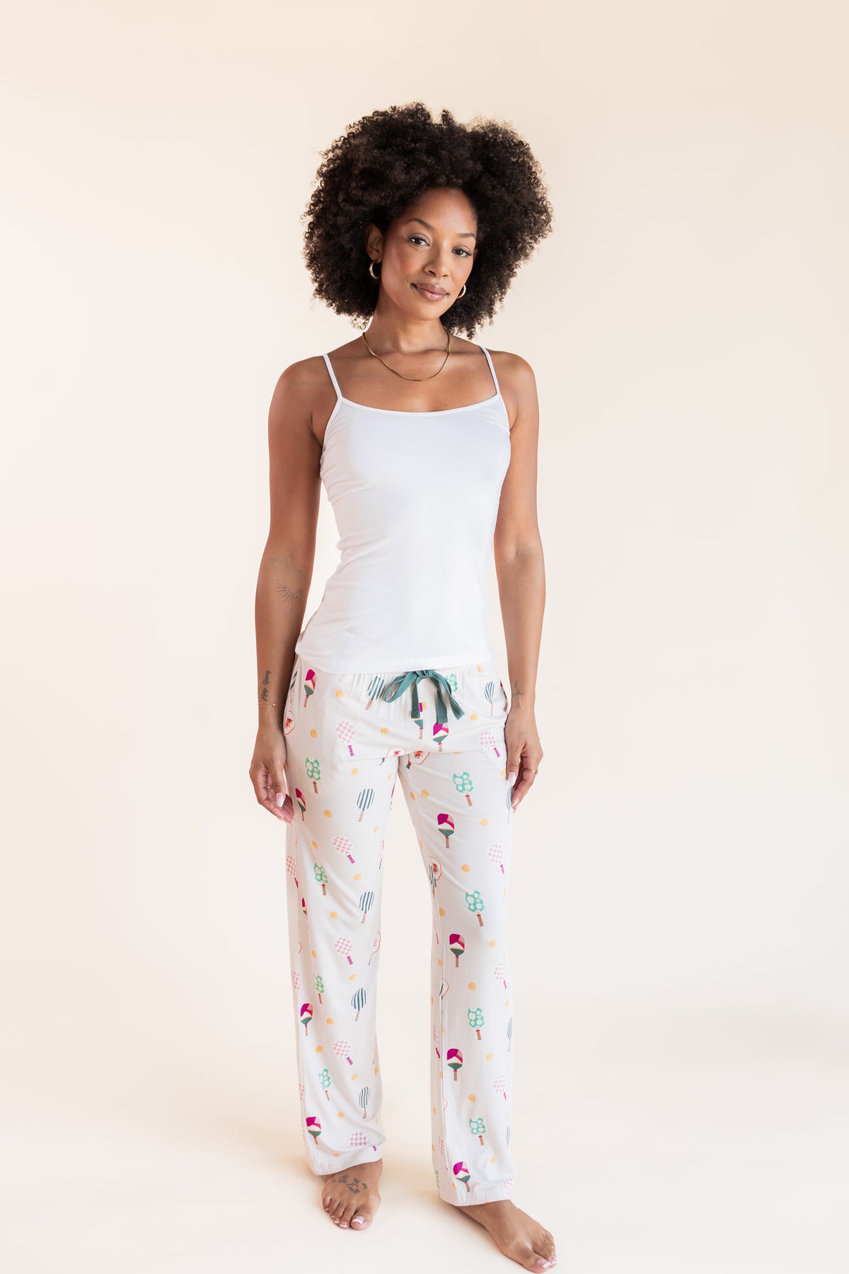 A woman modeling a two-piece pajama set consisting of a white camisole top and white pants with a colorful pickleball  print, set against a solid cream background.