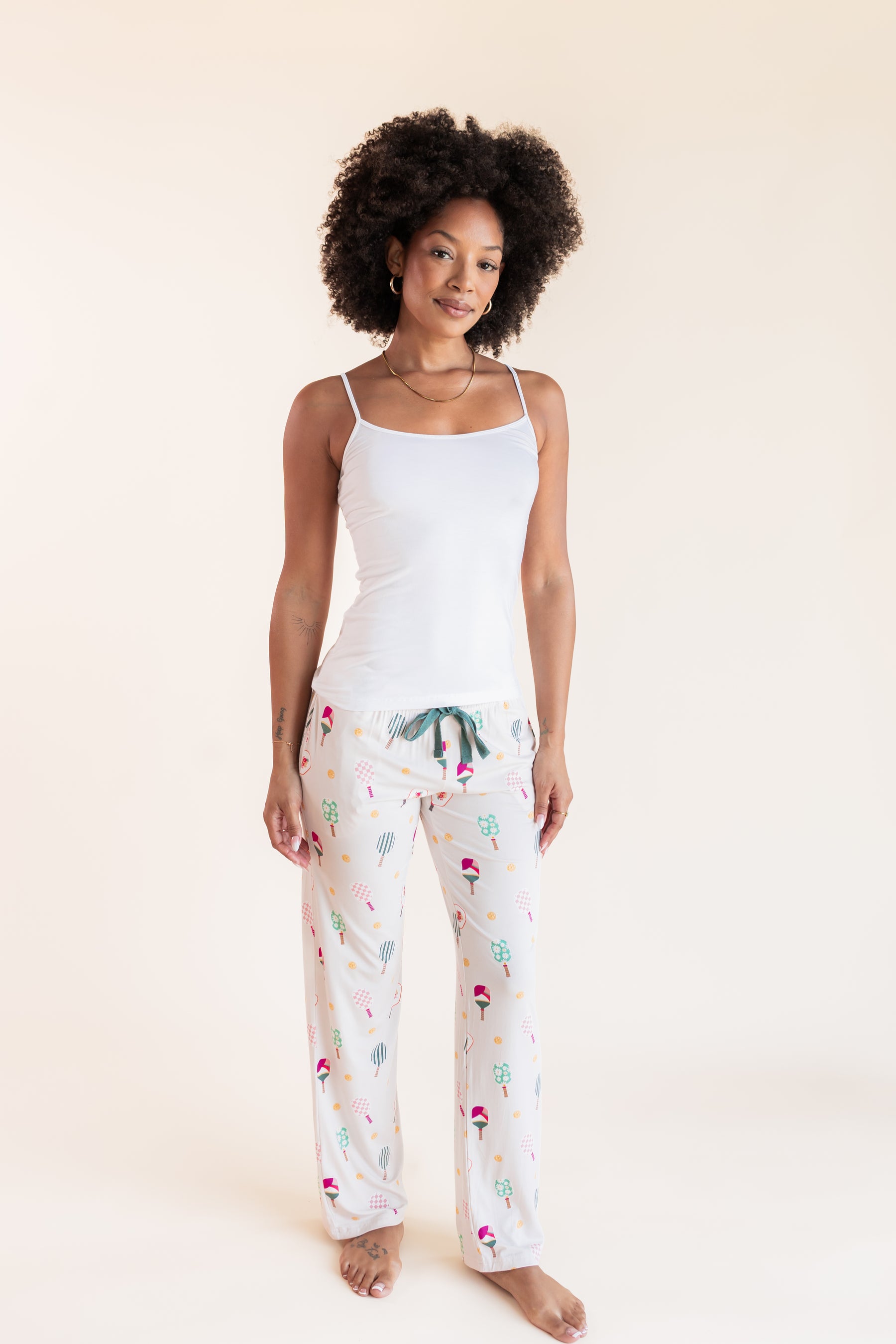 A woman modeling a two-piece pajama set consisting of a white camisole top and white pants with a colorful pickleball  print, set against a solid cream background.