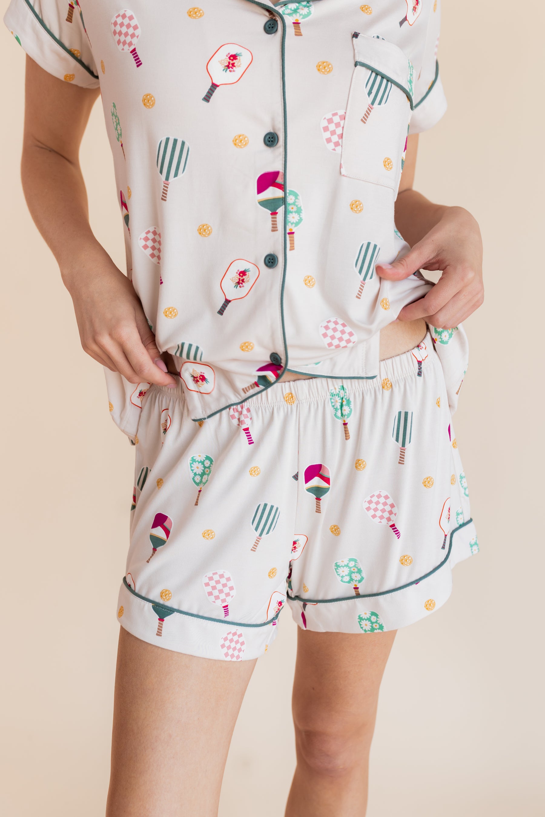 A person wearing a matching short-sleeve button-front pajama top and shorts set in a light cream color with a playful multicolor print of pickleball rackets