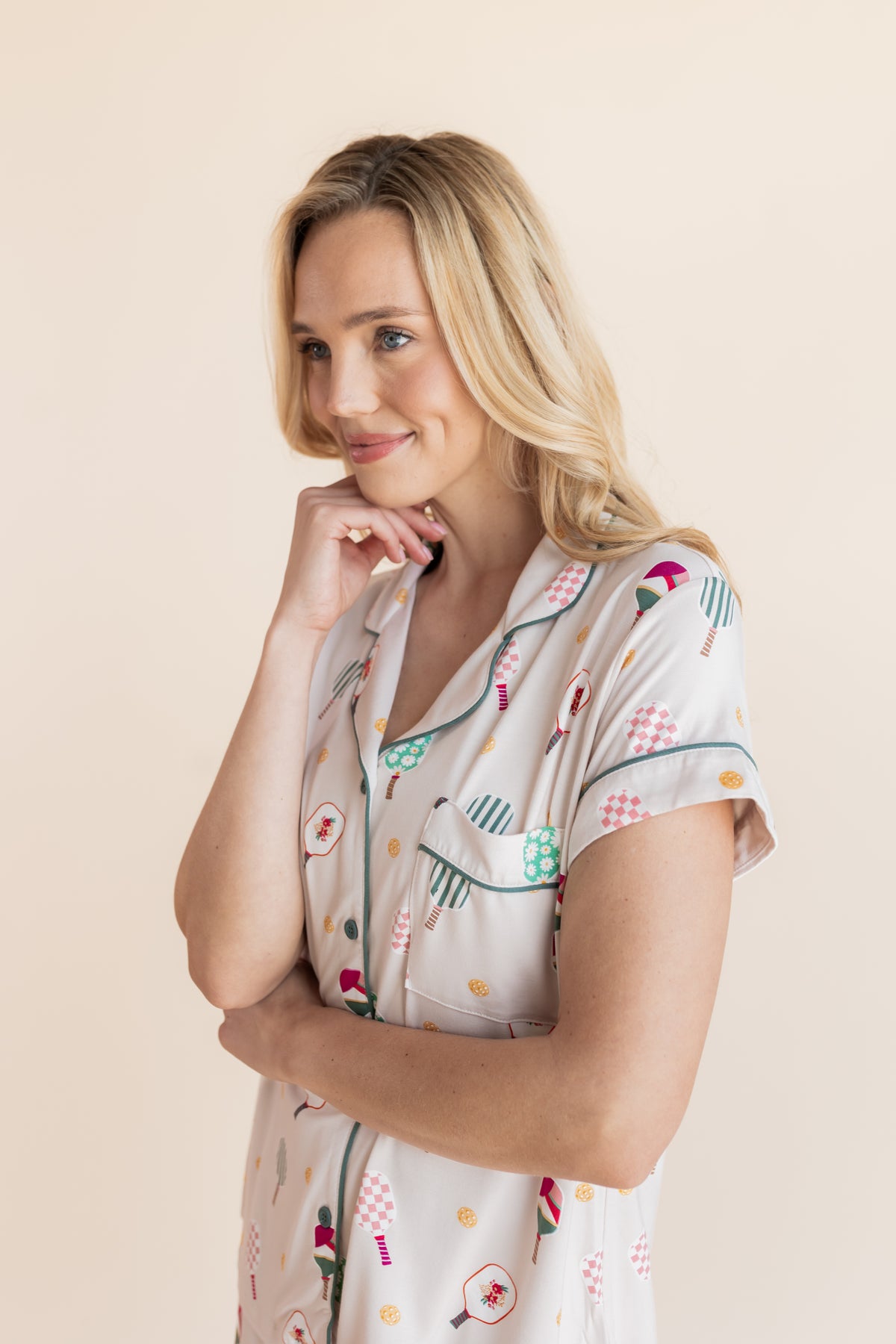 A blonde woman wearing a short-sleeve pajama top with a colorful pickleball-themed print, posing against a solid light beige background.