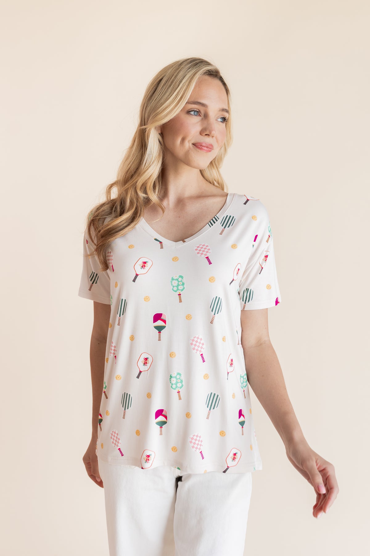 A woman modeling a white short-sleeve V-neck top with a colorful pickleball print, paired with white pants, against a solid beige background.
