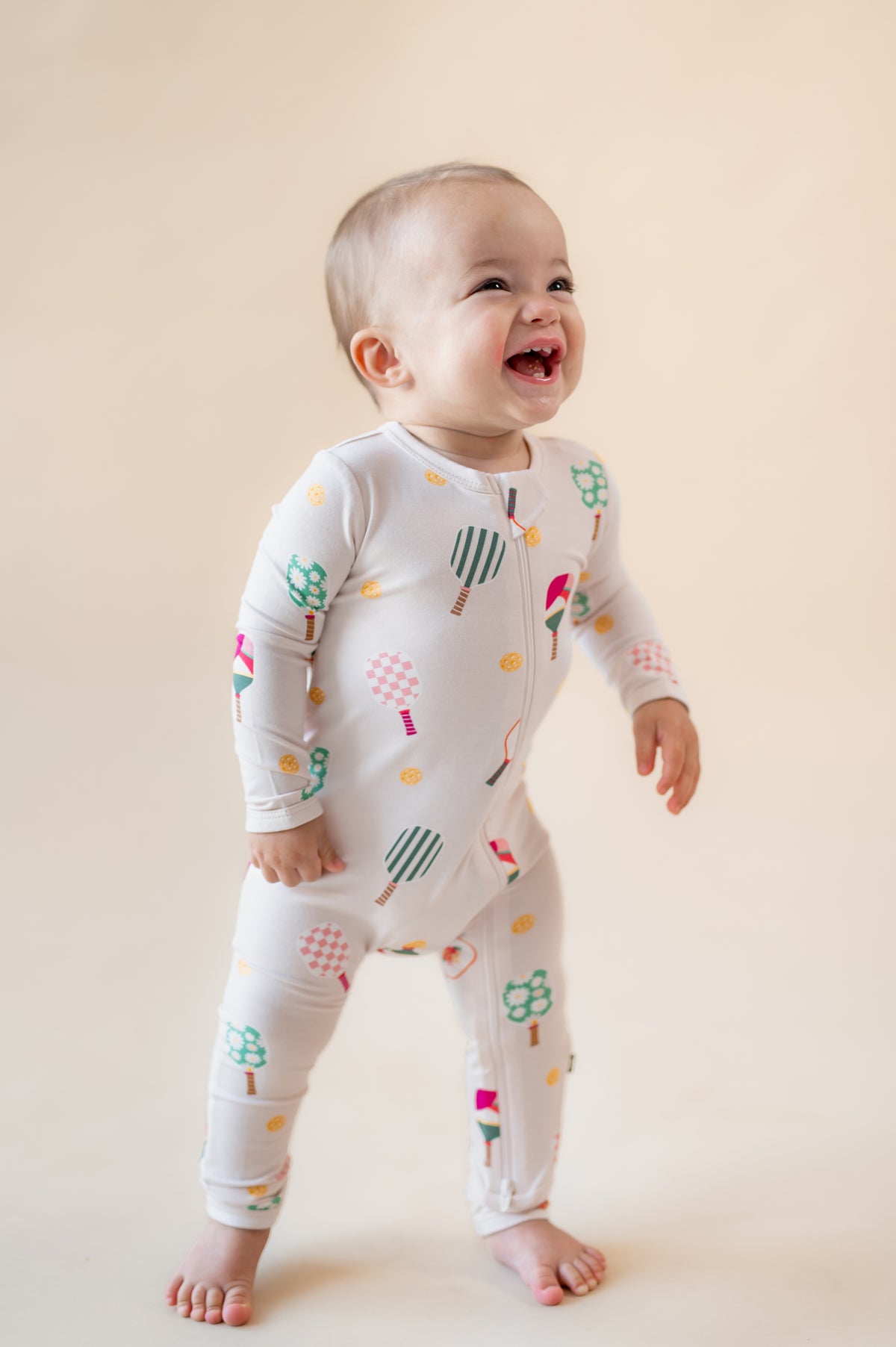 Baby smiling in Zippered Romper in Pickleball print on a beige background
