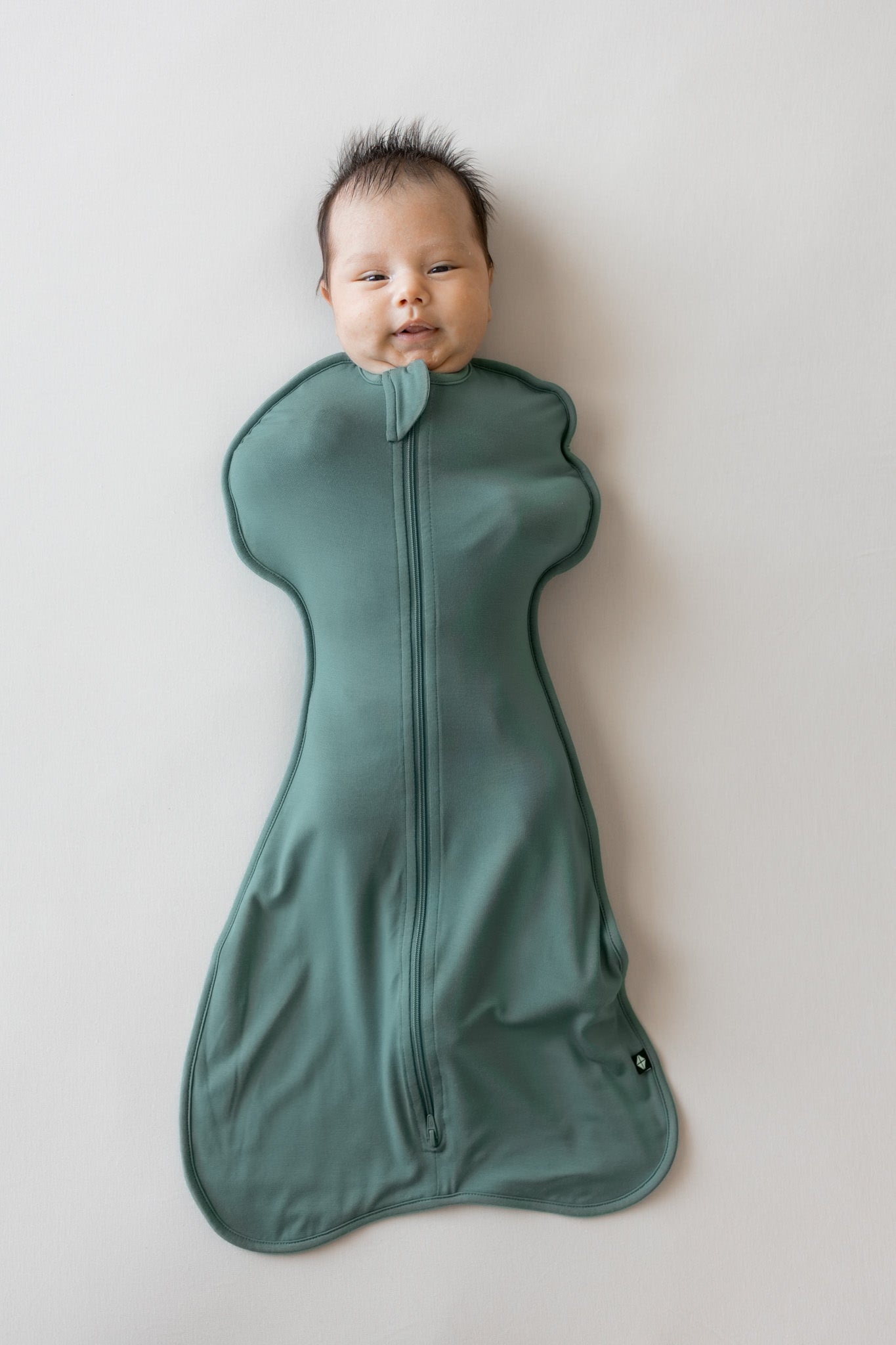 A baby lying on their back wrapped in a sage green zippered swaddle bag against a plain light gray background.