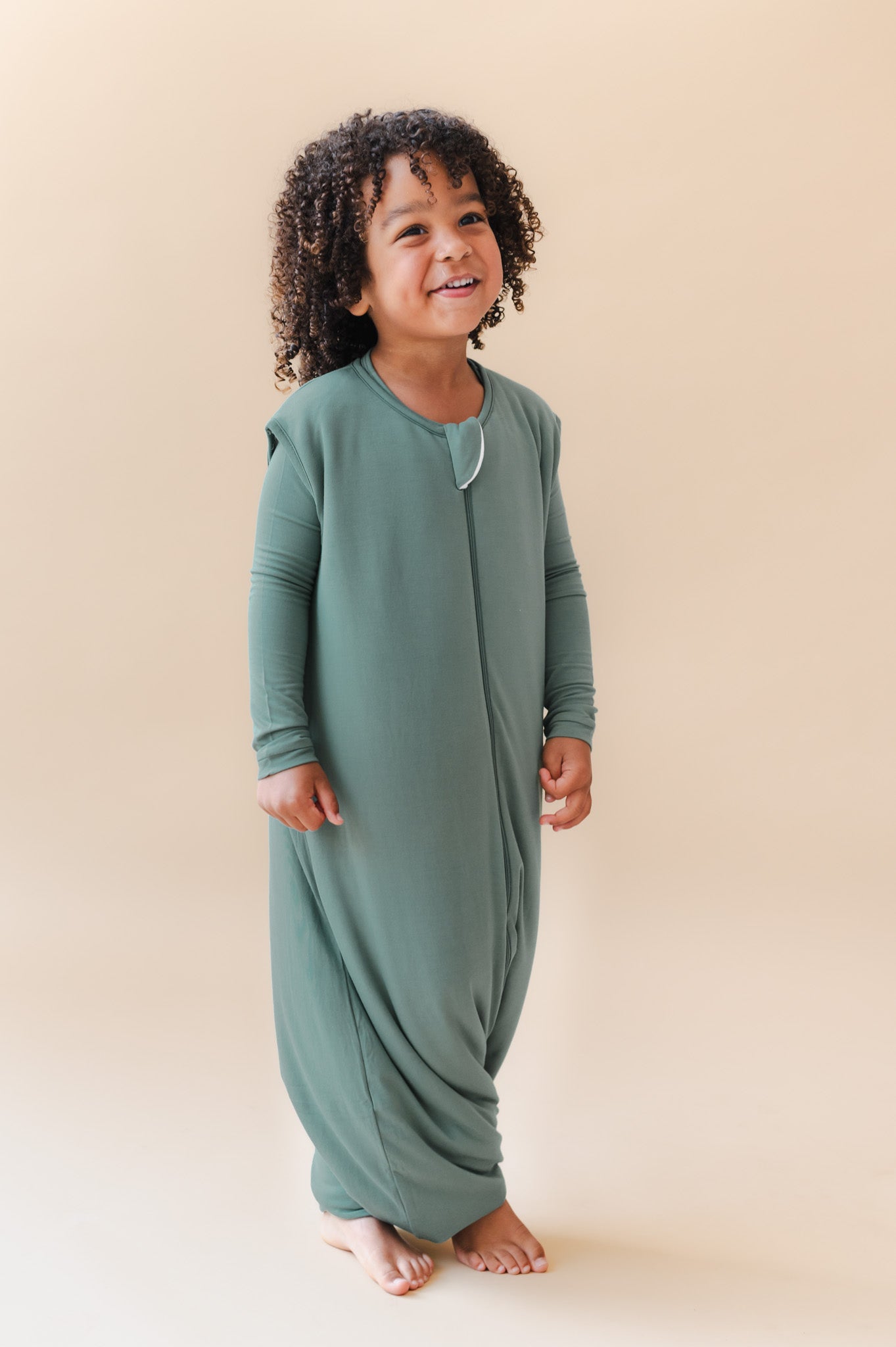 A smiling toddlerstands barefoot against a warm beige studio background, wearing a sage green footless sleep sack 