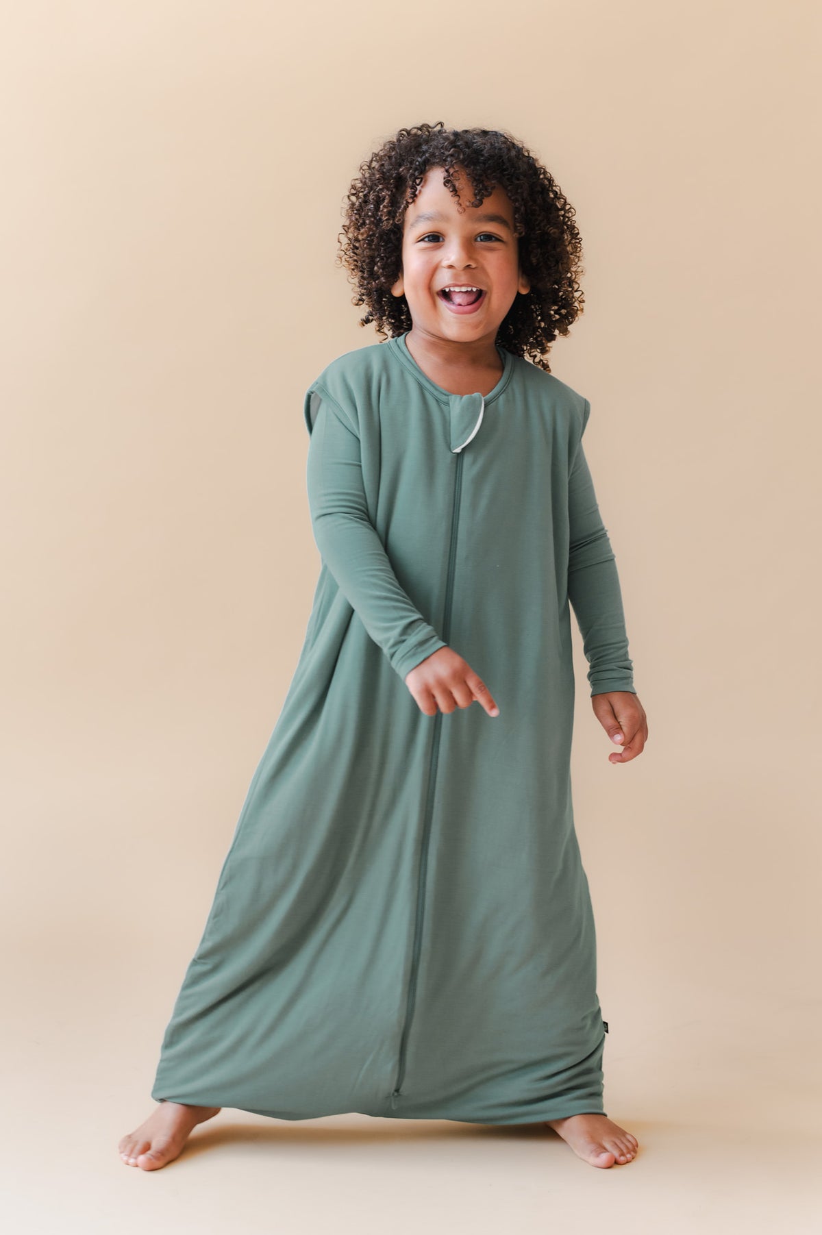 A smiling young child stands barefoot against a plain warm beige studio background, wearing a long-sleeve sleep sack in muted sage green.