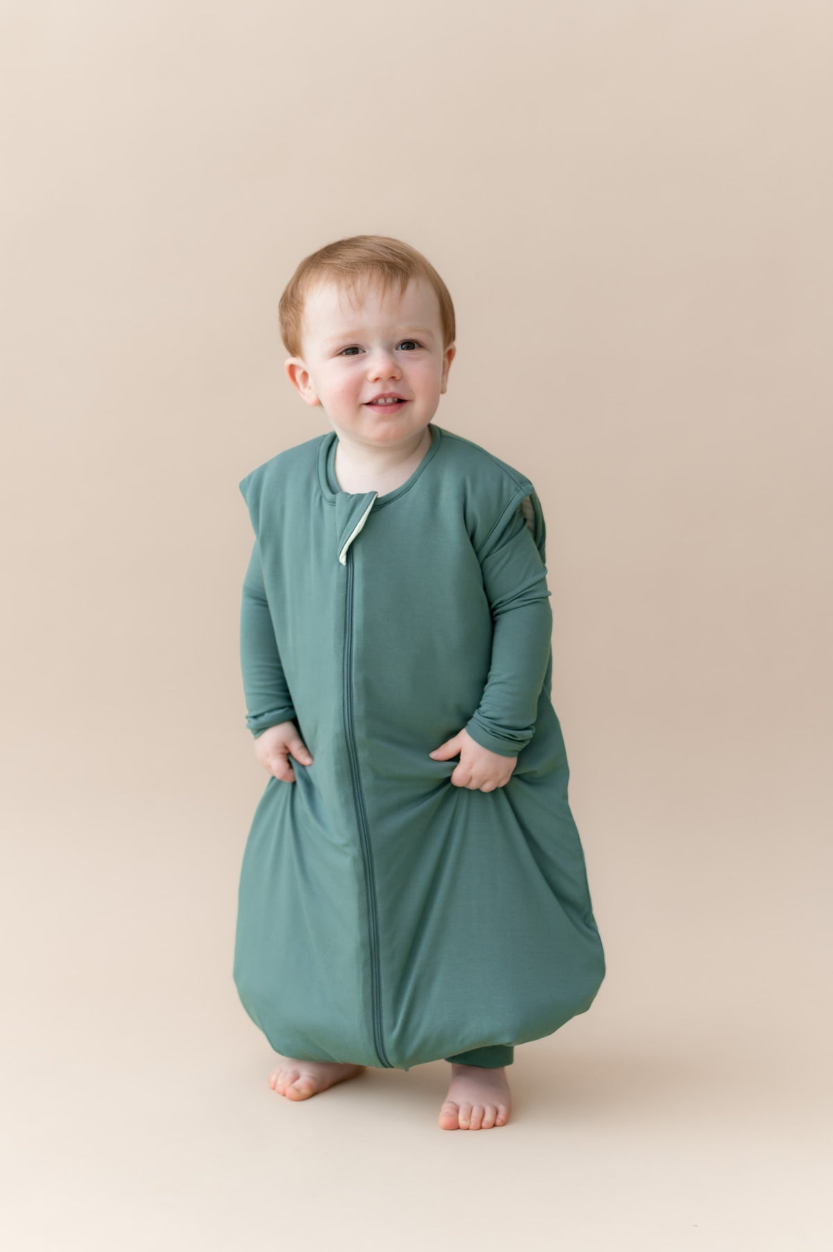 A toddler wearing a sage green zip-up sleeping bag walker, standing against a solid beige studio background.