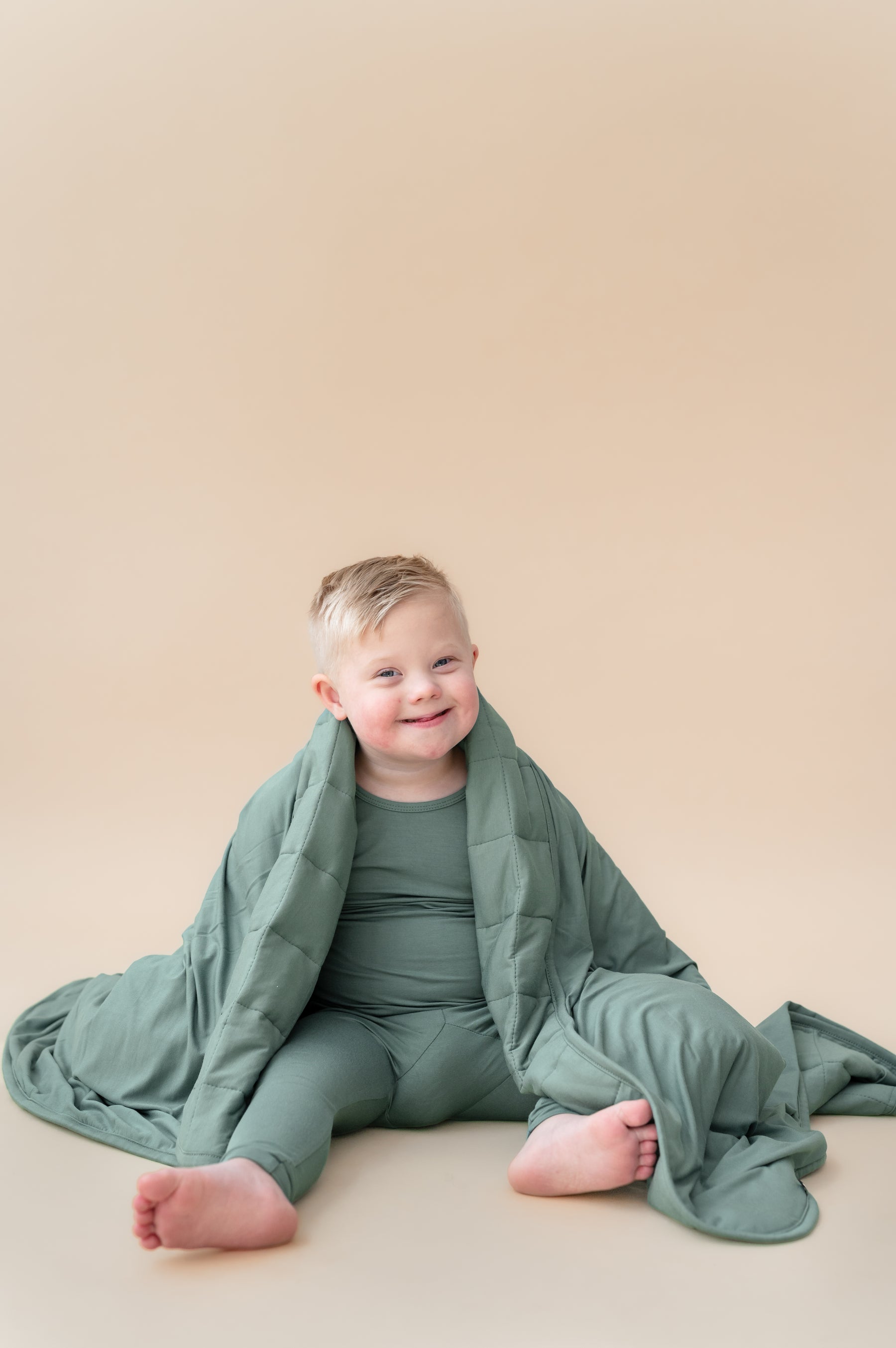 Toddler sitting with Toddler Blanket in Pine 2.5 wrapped over his shoulder wearing similar color long sleeve pajama set on a beige background