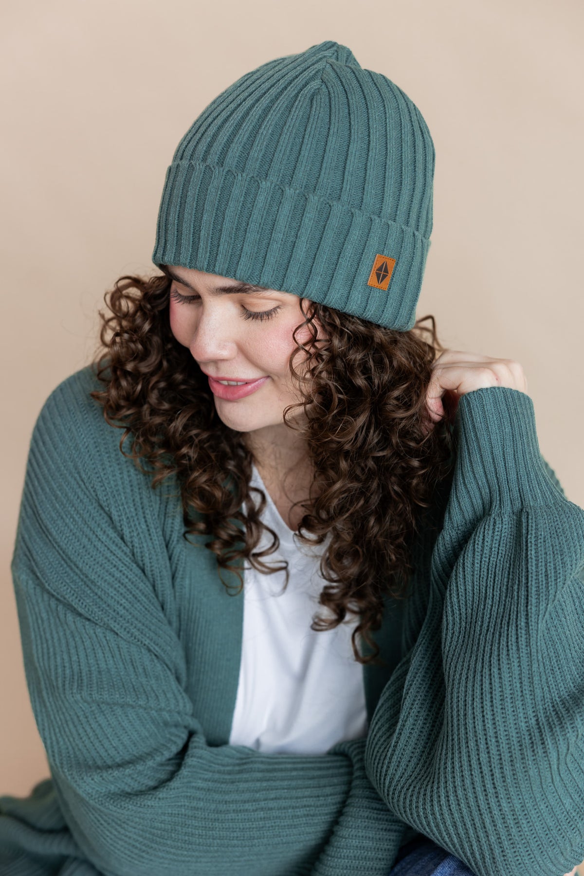 Modal posing in Chunky Knit Adult Ribbed Beanie in Pine over white T-shirt and Chunky knit cardigan in Pine on a beige background