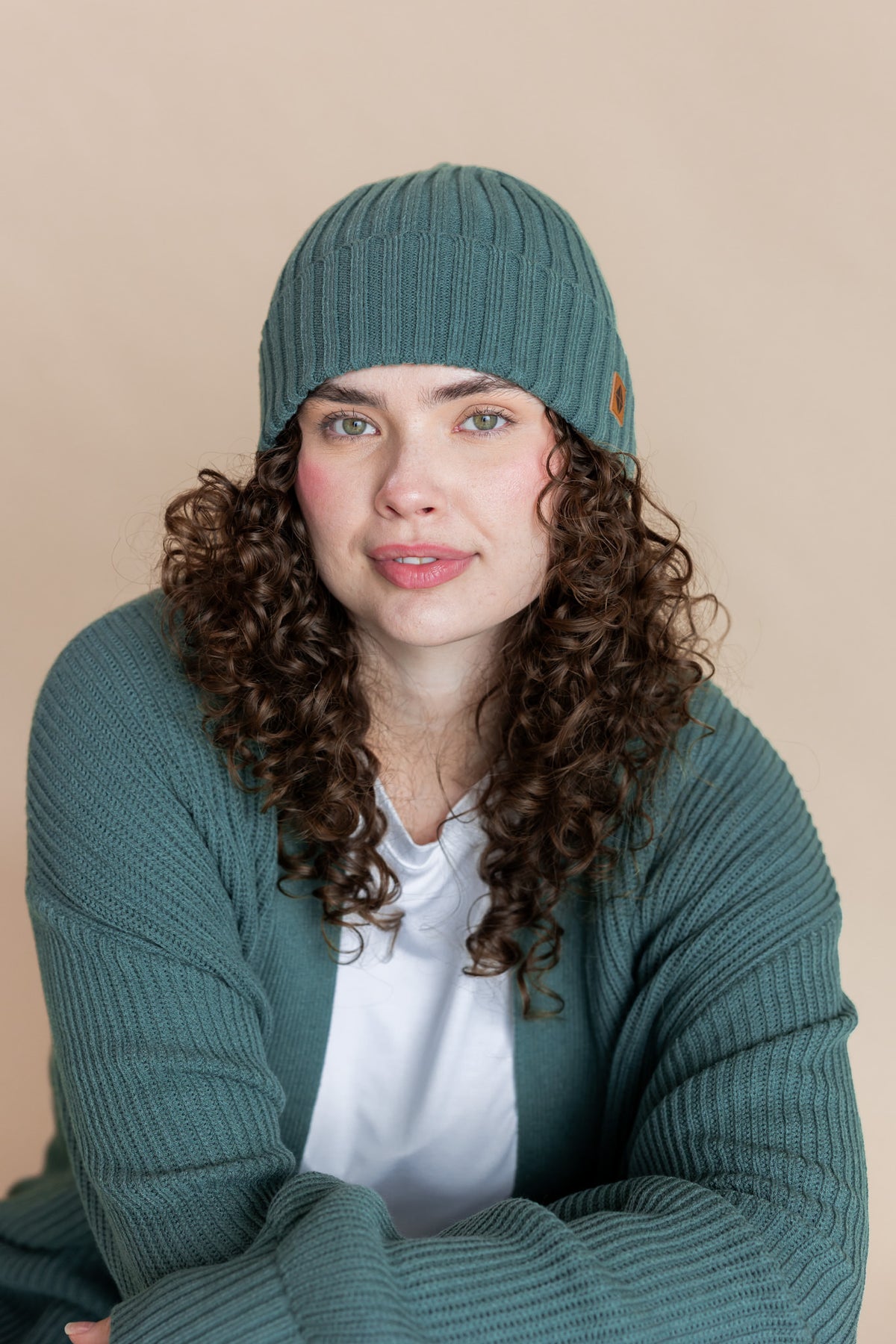 Modal smiling in Chunky Knit Adult Ribbed Beanie in Pine over white T-shirt and Chunky knit cardigan in Pine on a beige background