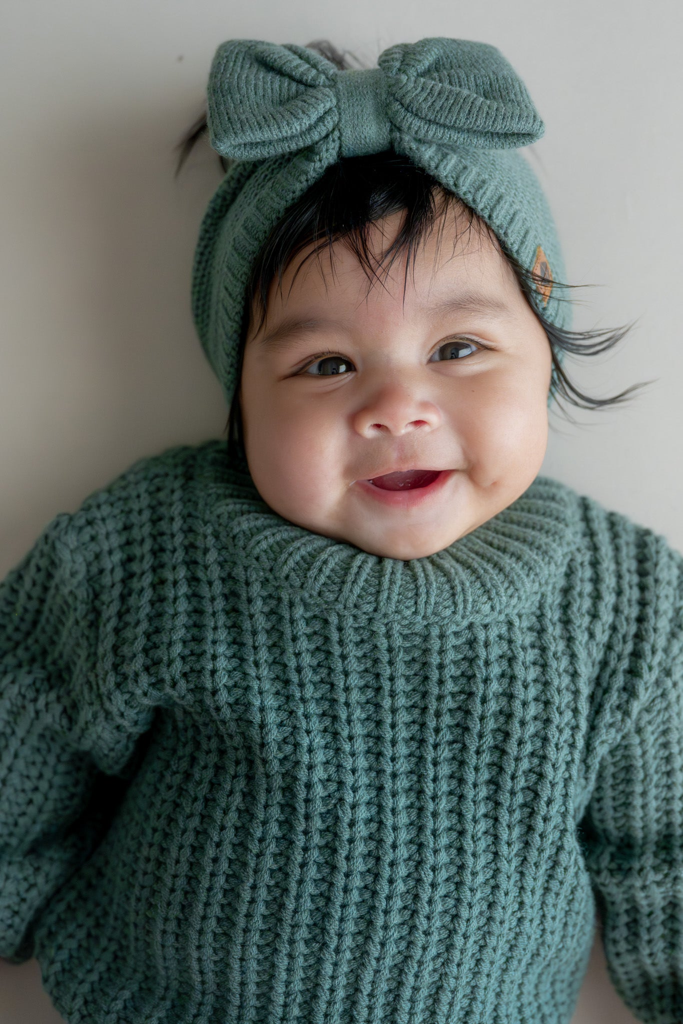 Baby wearing Chunky Knit Bow Headband in Pine over Chunky knit sweater in pine on a beige background