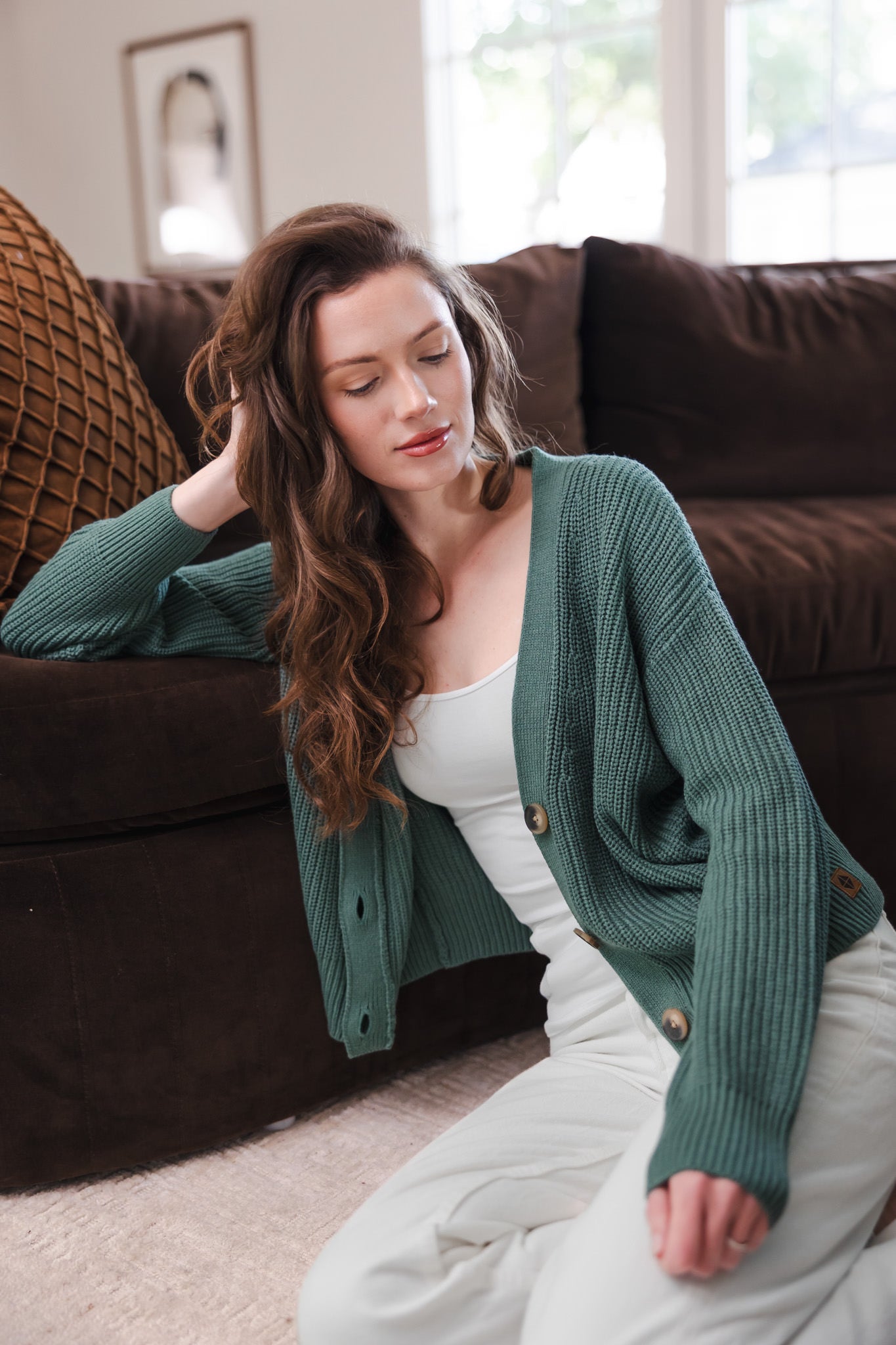 Modal sitting against a brown couch while wearing Chunky Knit Women's Cropped Cardigan in Pine over white camisole and white pants