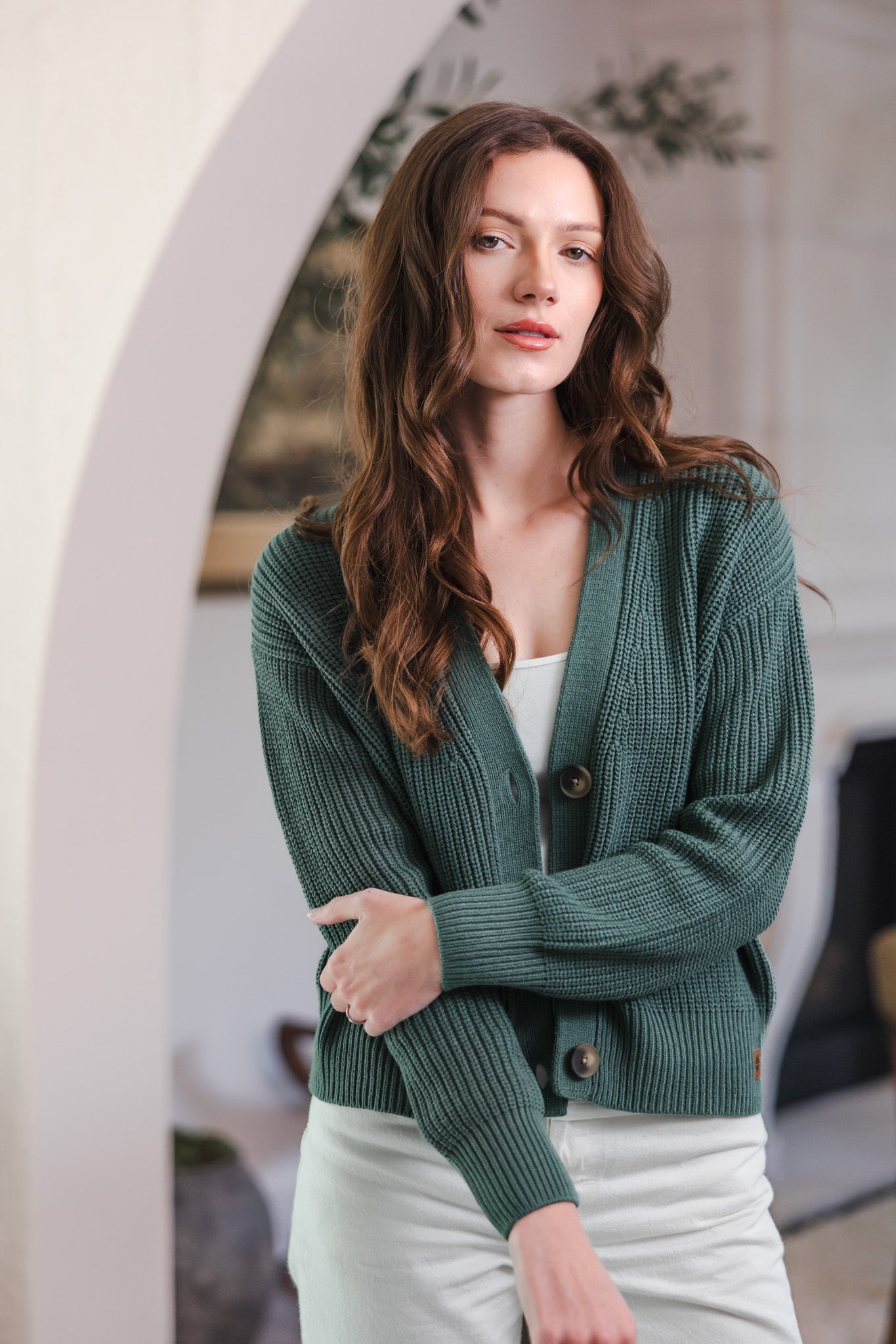 Modal standing in living room while posing in Chunky Knit Women's Cropped Cardigan in Pine over white pants