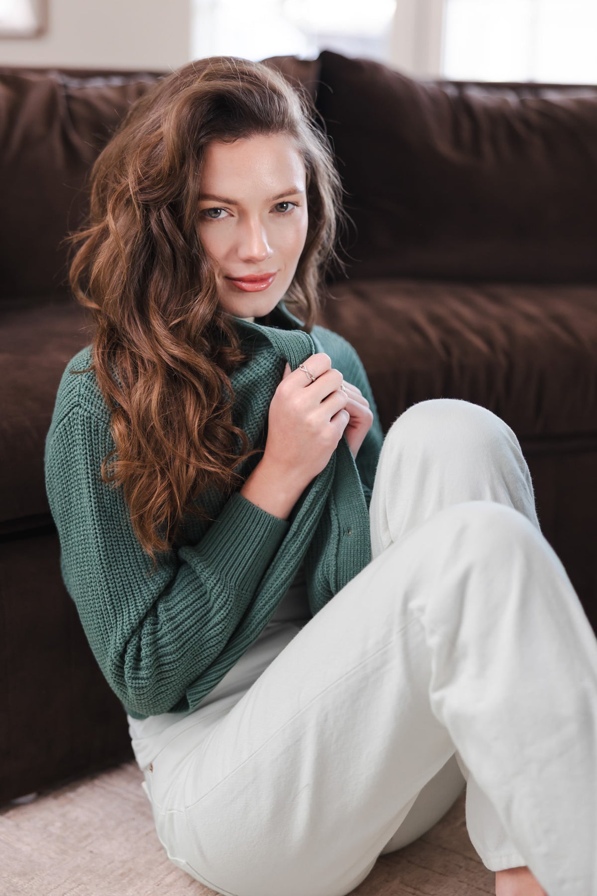 Modal sitting in a living room against a brow sofa while wearing Chunky Knit Women's Cropped Cardigan in Pine over white pants