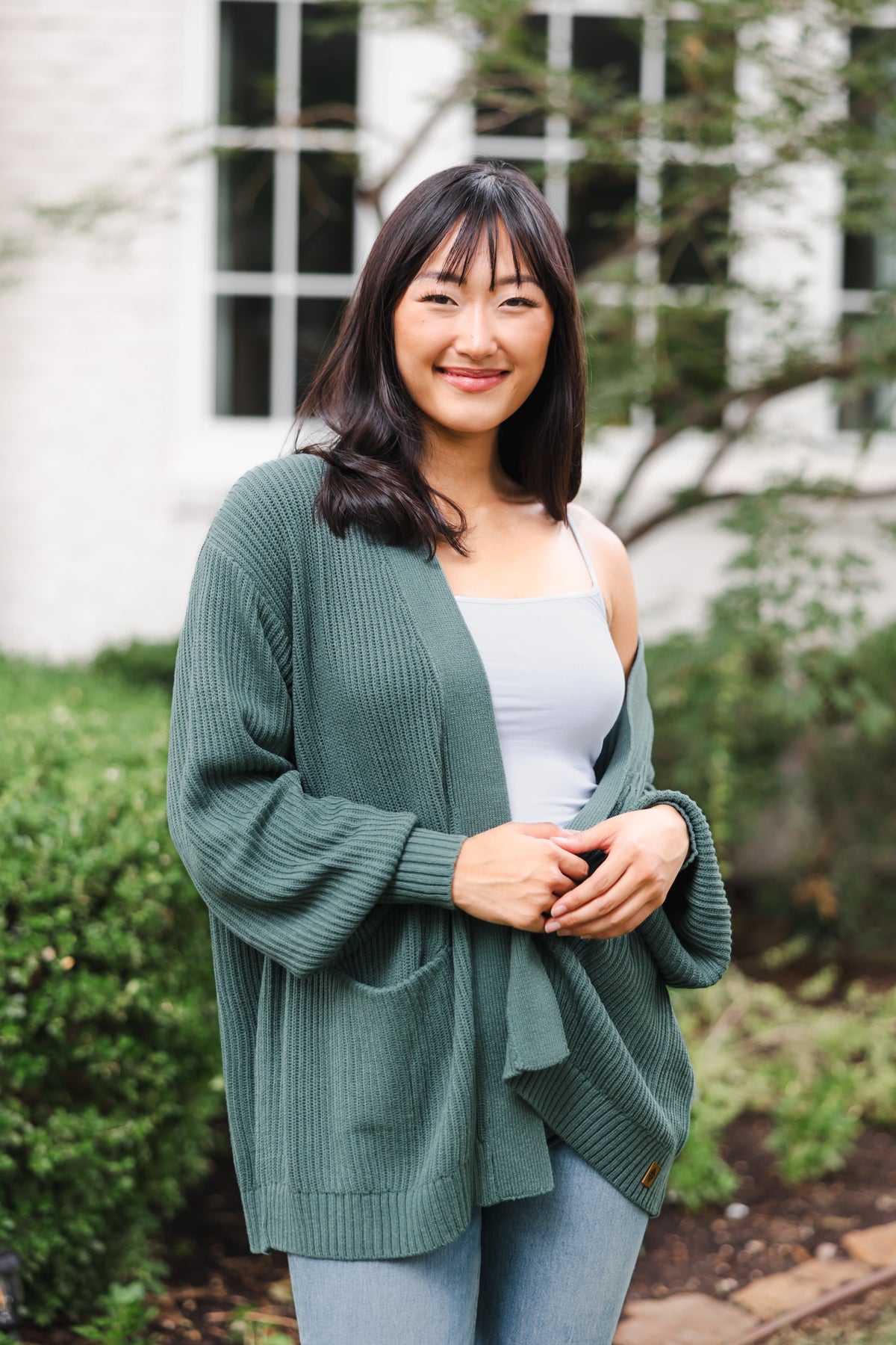 Modal smiling outdoors wearing Chunky knit oversized cardigan in pine over white camisole and blue jeans