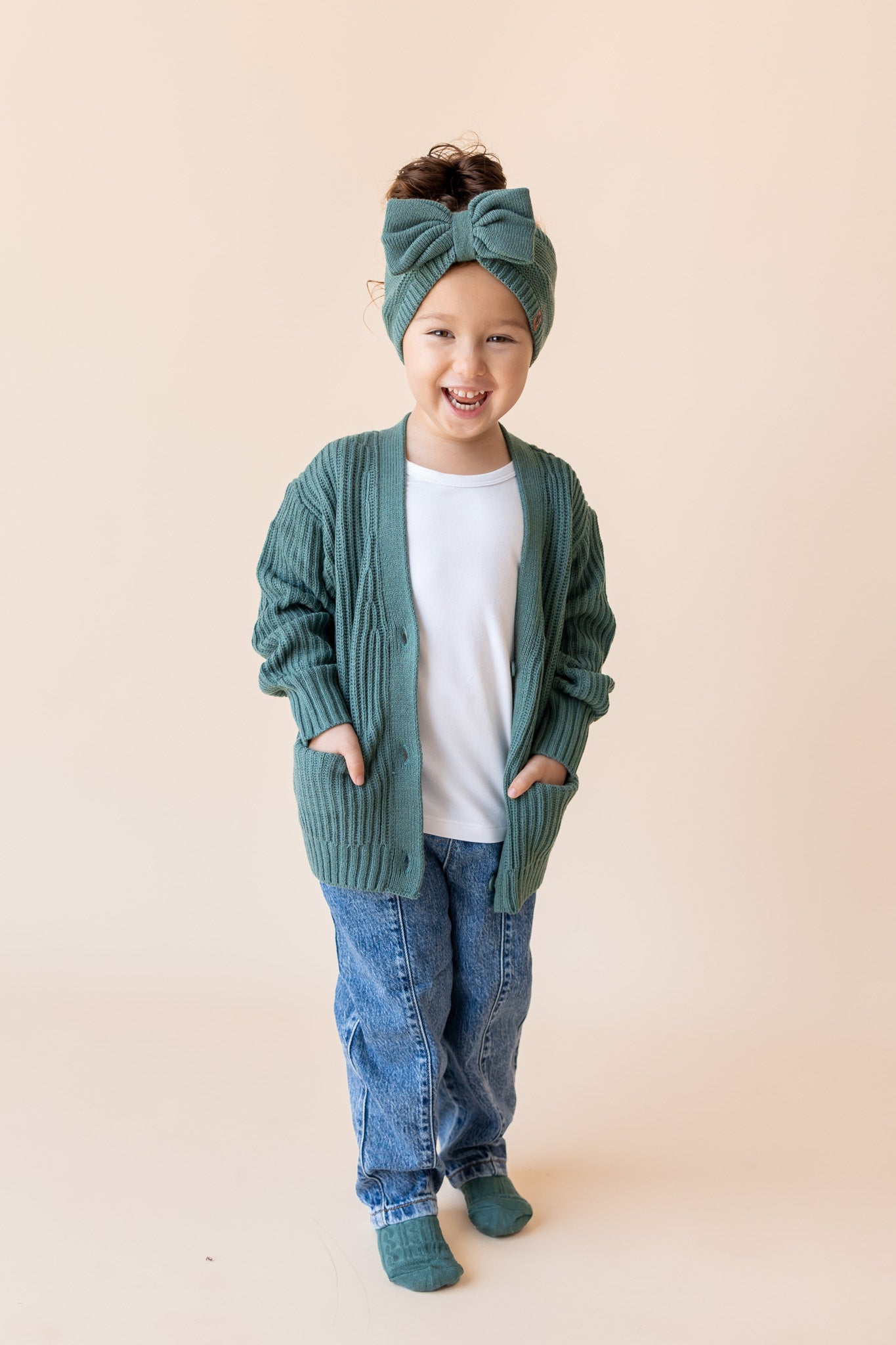 Girl wearing Chunky knit oversized cardigan, chunky knit bow headband in pine over white T-shirt and blue jeans on a beige background