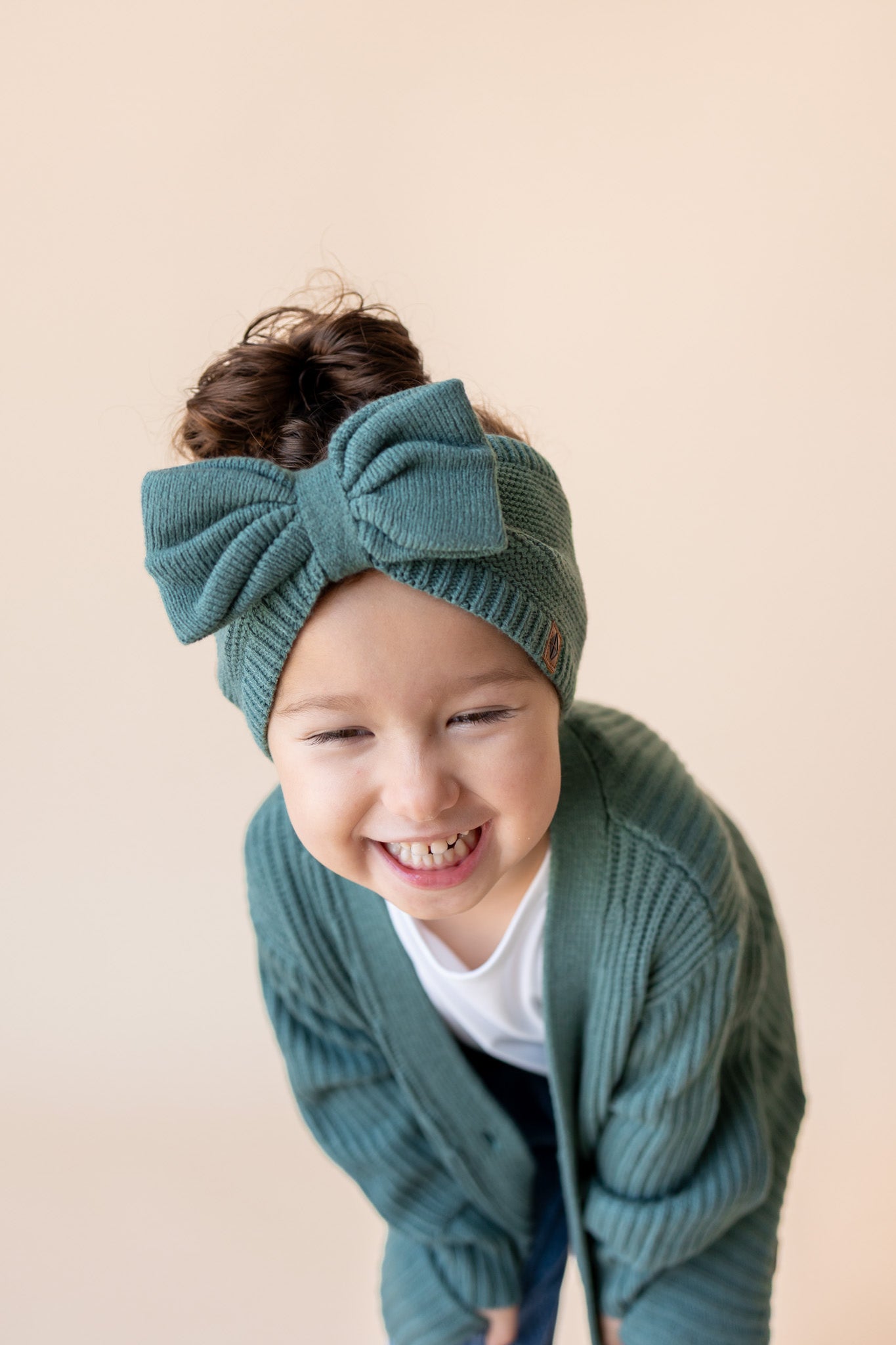 Modal smiling in Chunky Knit Bow Headband in Pine over white T-shirt and Chunky knit oversized cardigan in pine on a beige background
