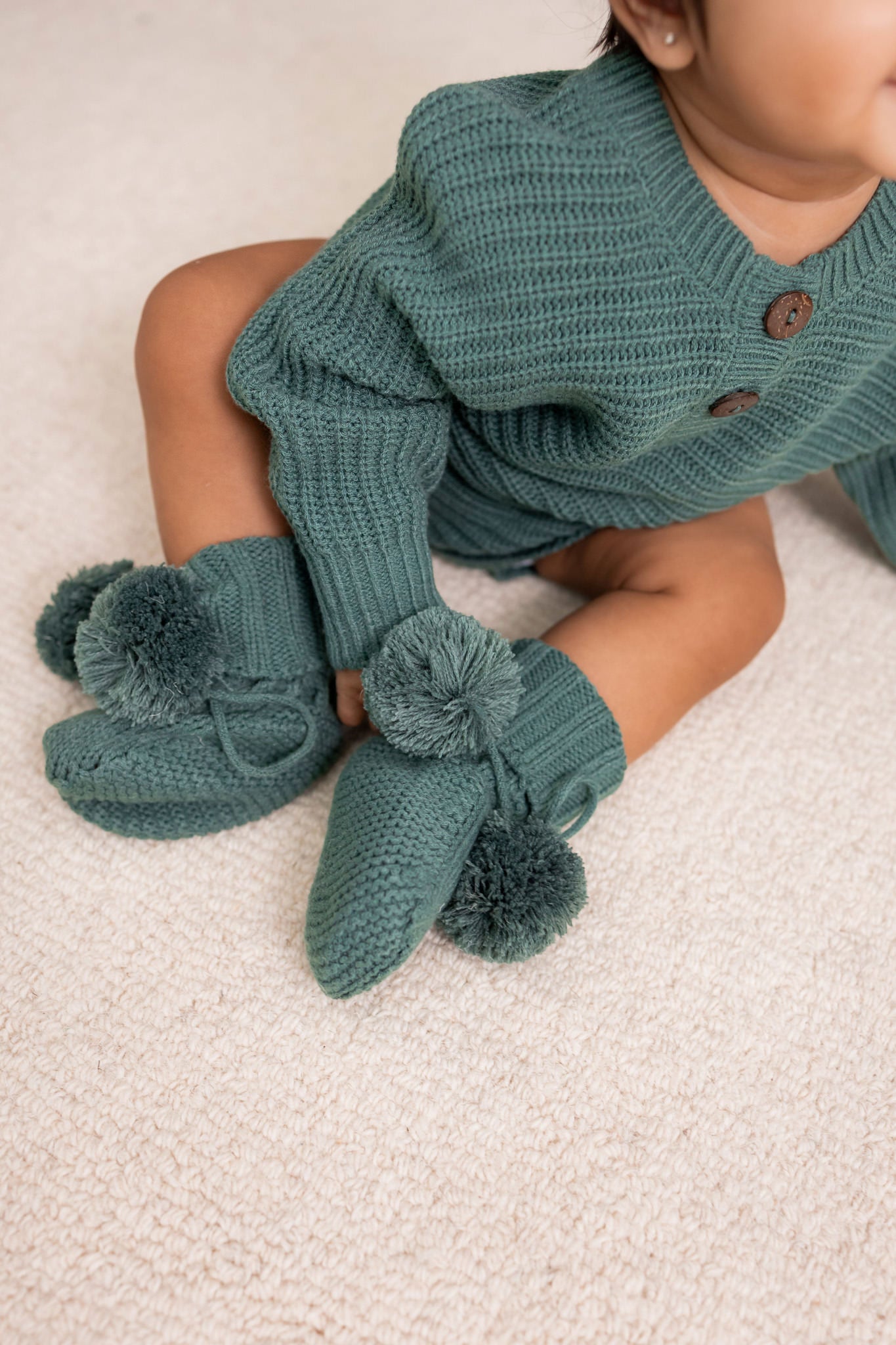 Baby wearing Knit Booties and chunky knit romper in Pine on a beige carpet