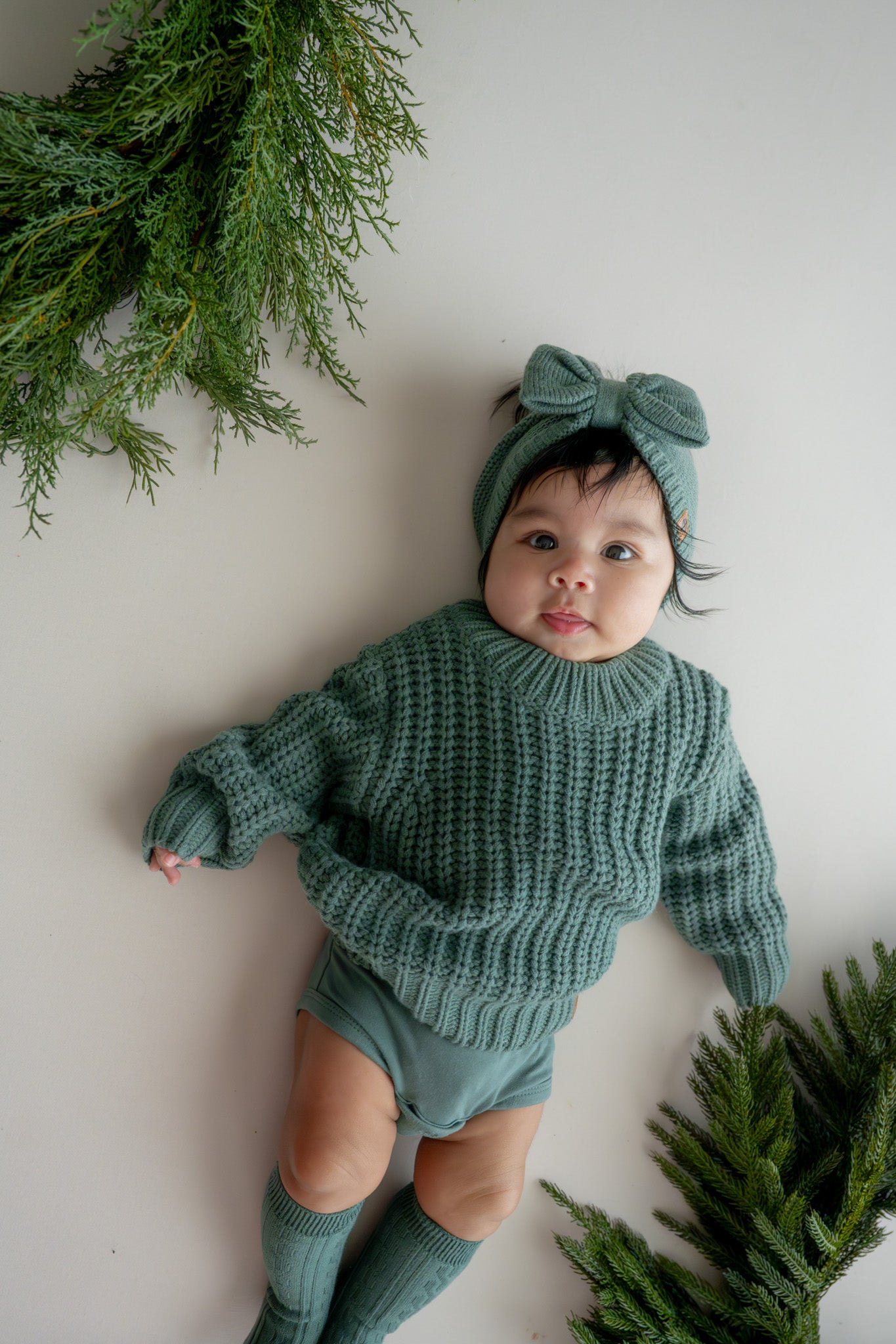 Baby wearing Chunky Knit Sweater,  chunky knit bow headband in Pine and socks in pine lying on a beige background 