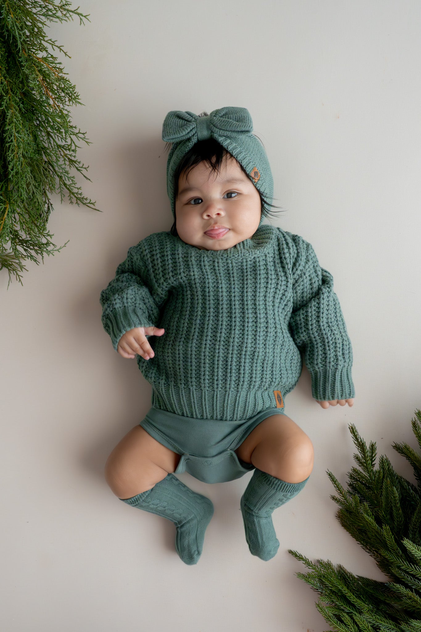 Baby wearing Chunky Knit Bow Headband in Pine over Chunky knit sweater in pine and pine knee high socks on a beige background
