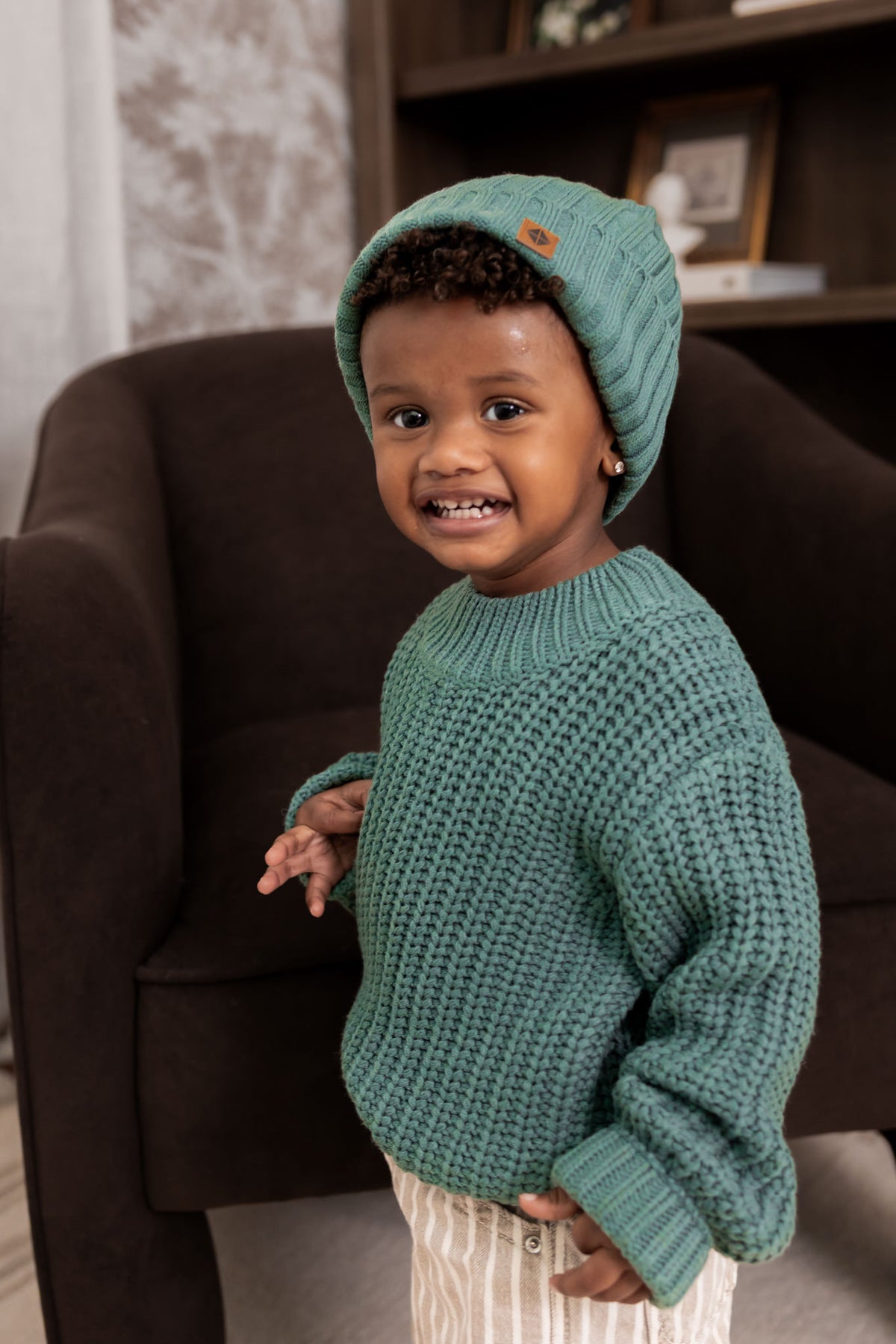 Boy wearing Chunky Knit Sweater and chunky knit ribbed beanie in Pine over beige pants standing in a living room