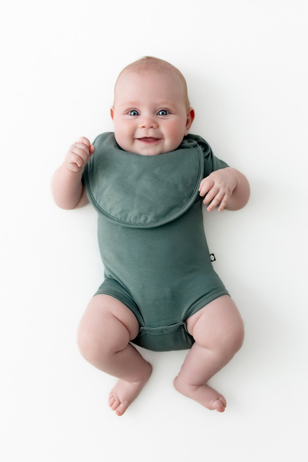 Baby smiling in Cushy Bib in Pine over similar color bodysuit on a white background
