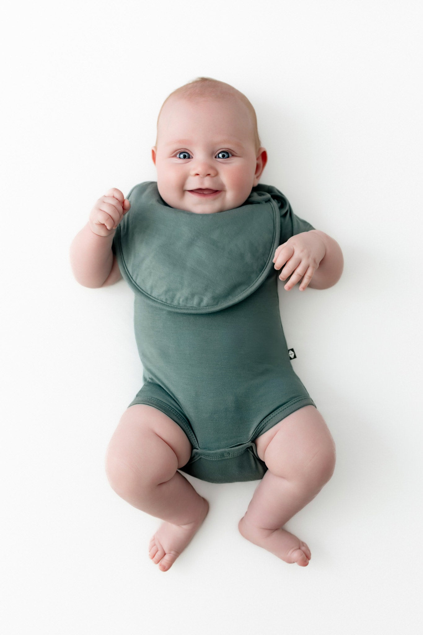 Baby smiling in Cushy Bib in Pine over similar color bodysuit on a white background