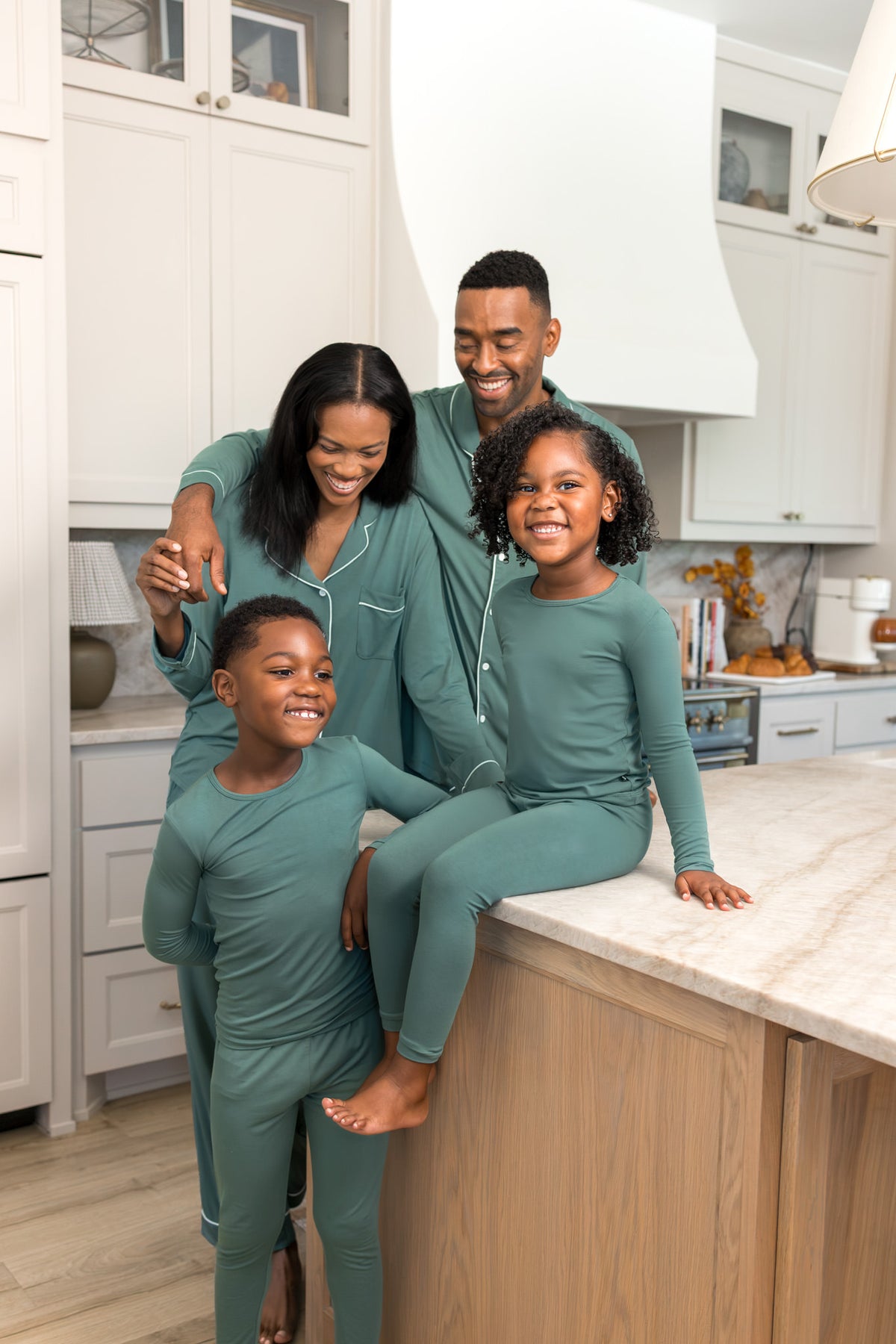 A family of four wearing matching sage green long-sleeve pajama sets poses together in a bright, modern kitchen. The set includes a matching top and pants, shown on a man, a woman, a young boy, and a young girl sitting on a kitchen island.