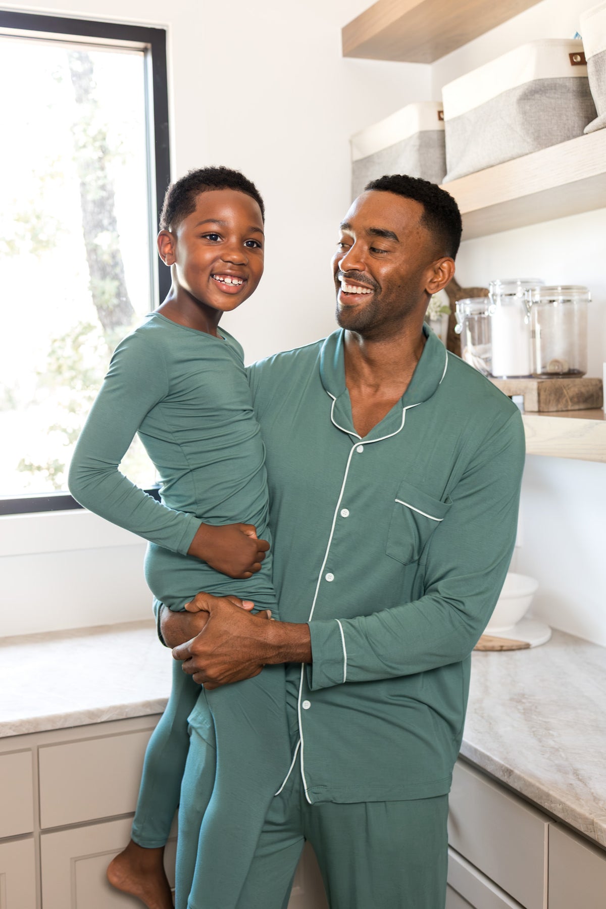 A man and a young boy wearing matching sage green pajamas with white piping, standing together in a bright kitchen setting.
