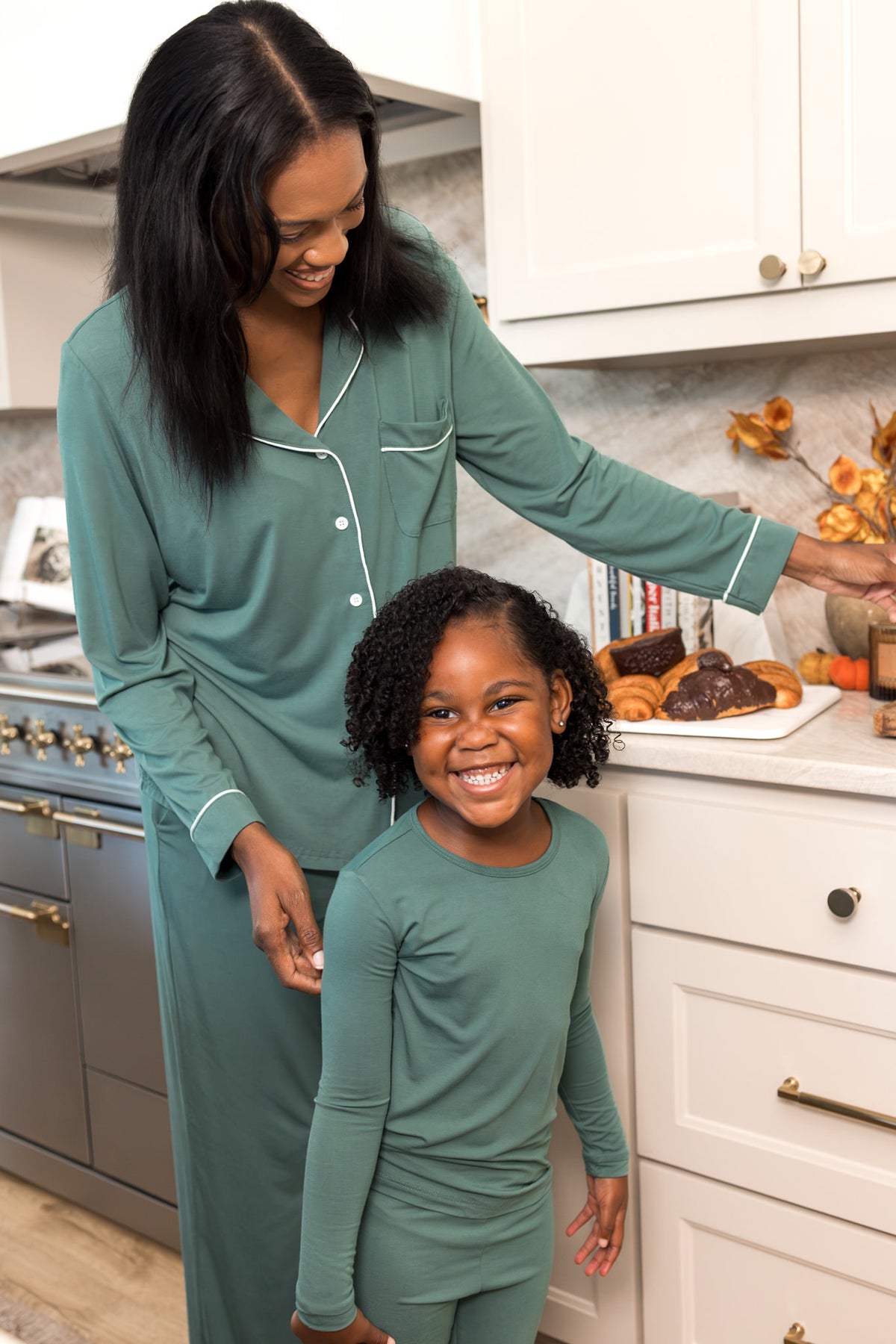 A woman and a young girl wearing matching sage green long-sleeve pajamas with white piping, standing in a bright kitchen.