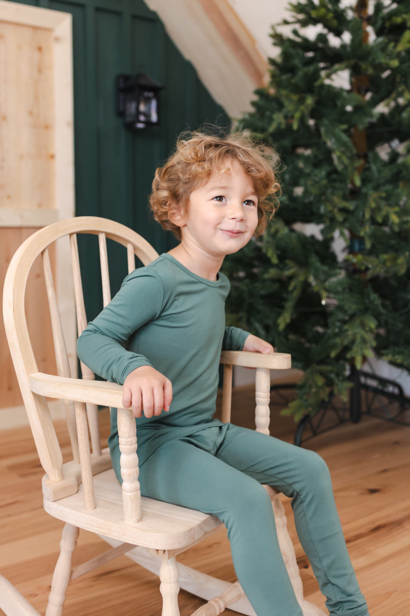 A young child wearing a matching sage green long-sleeve pajama set, sitting on a light wood rocking chair in a cozy room with a wooden floor and a Christmas tree in the background.