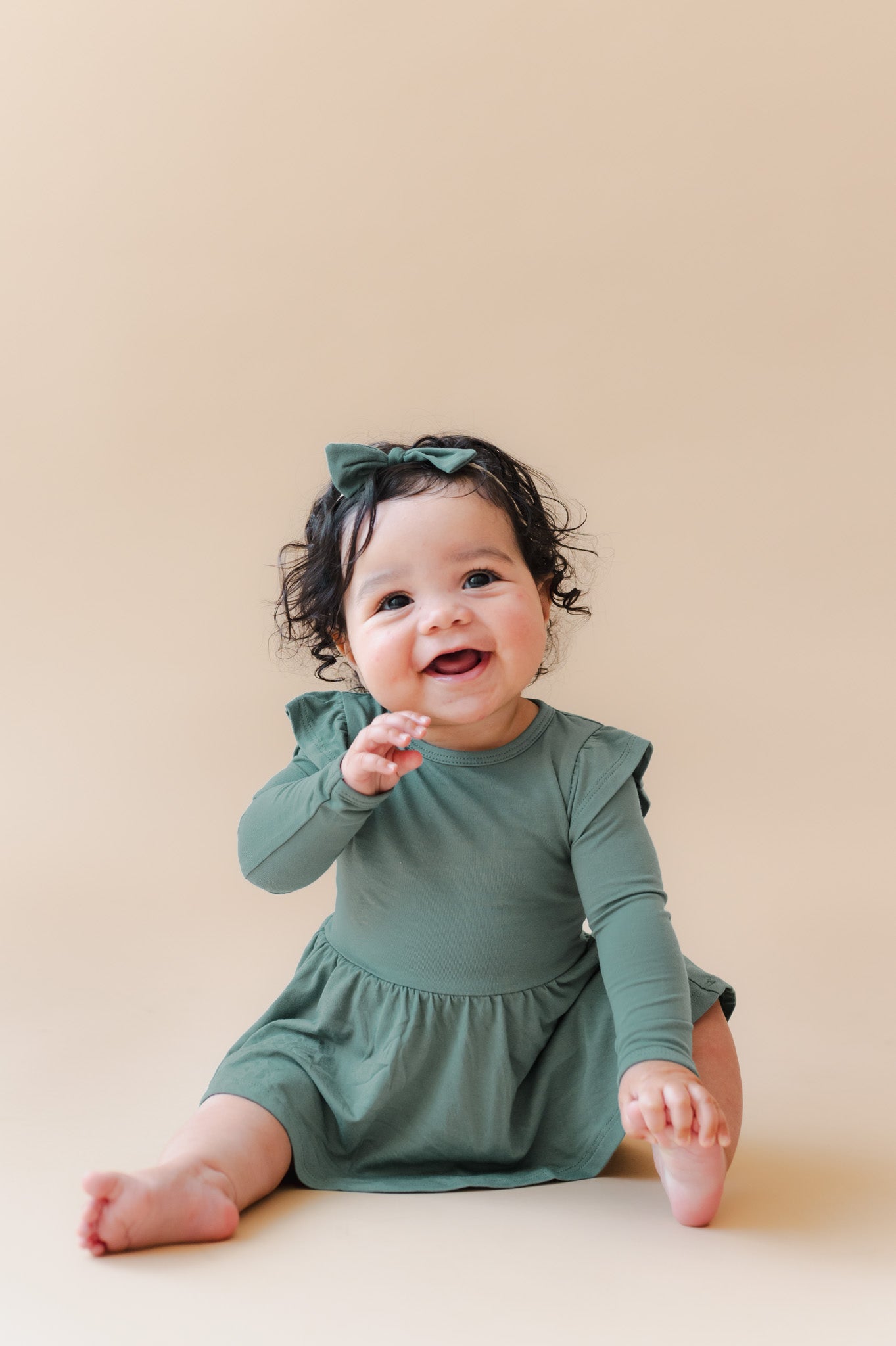 A smiling baby girl sitting on a beige studio floor, wearing a sage green long-sleeve bodysuit dress with ruffled shoulders and a matching green bow headband.