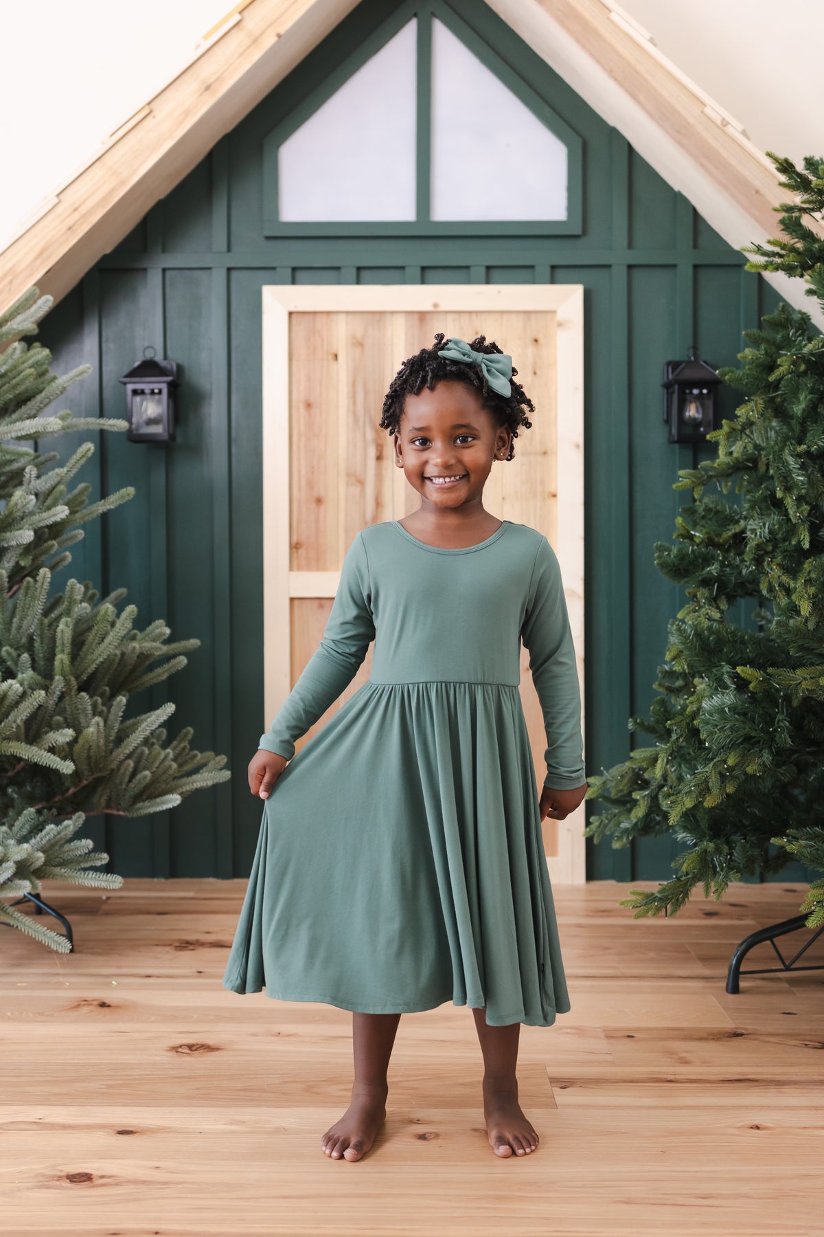 Girl wearing Long Sleeve Twirl Dress in Pine standing in front of a beige door