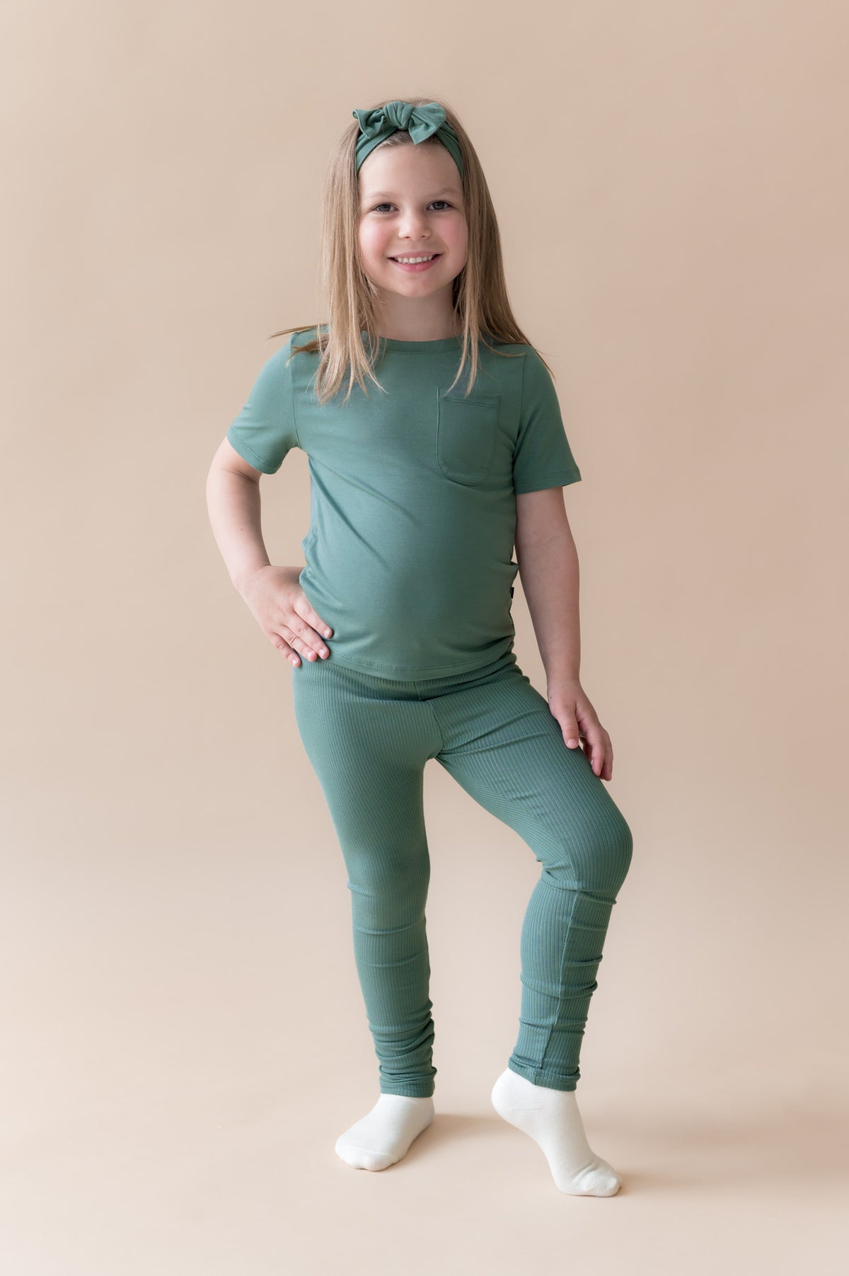 Girl wearing Ribbed Leggings and knotted bow headband in Pine on a beige background
