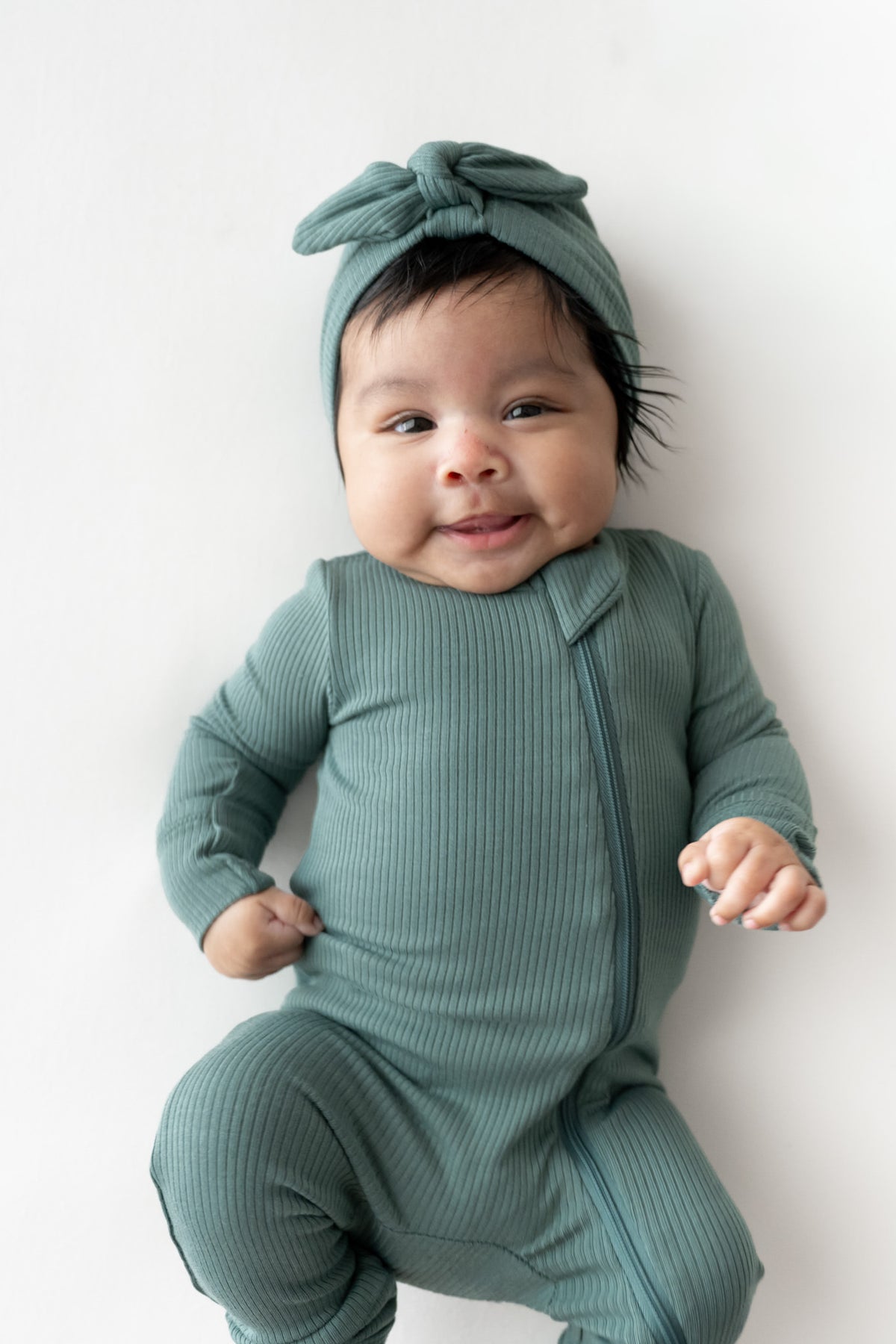 A baby wearing a ribbed sage green footed romper and matching bow headband, lying on a white background.