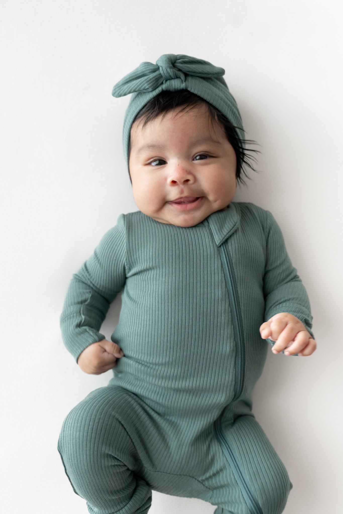 A baby wearing a ribbed sage green footed romper and matching bow headband, lying on a white background.