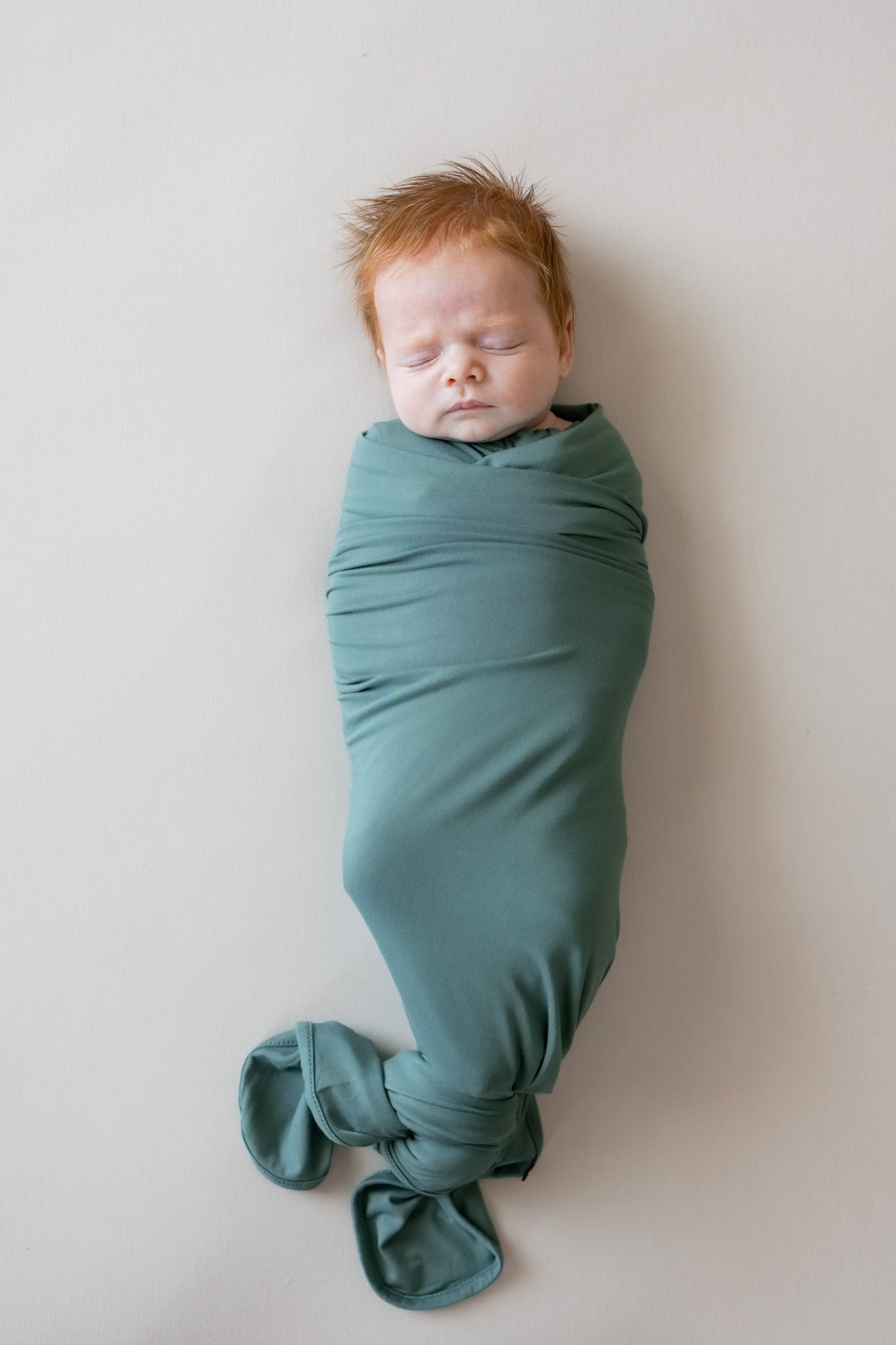 A sleeping newborn baby swaddled in a dusty green fabric wrap, lying on a neutral light beige background.