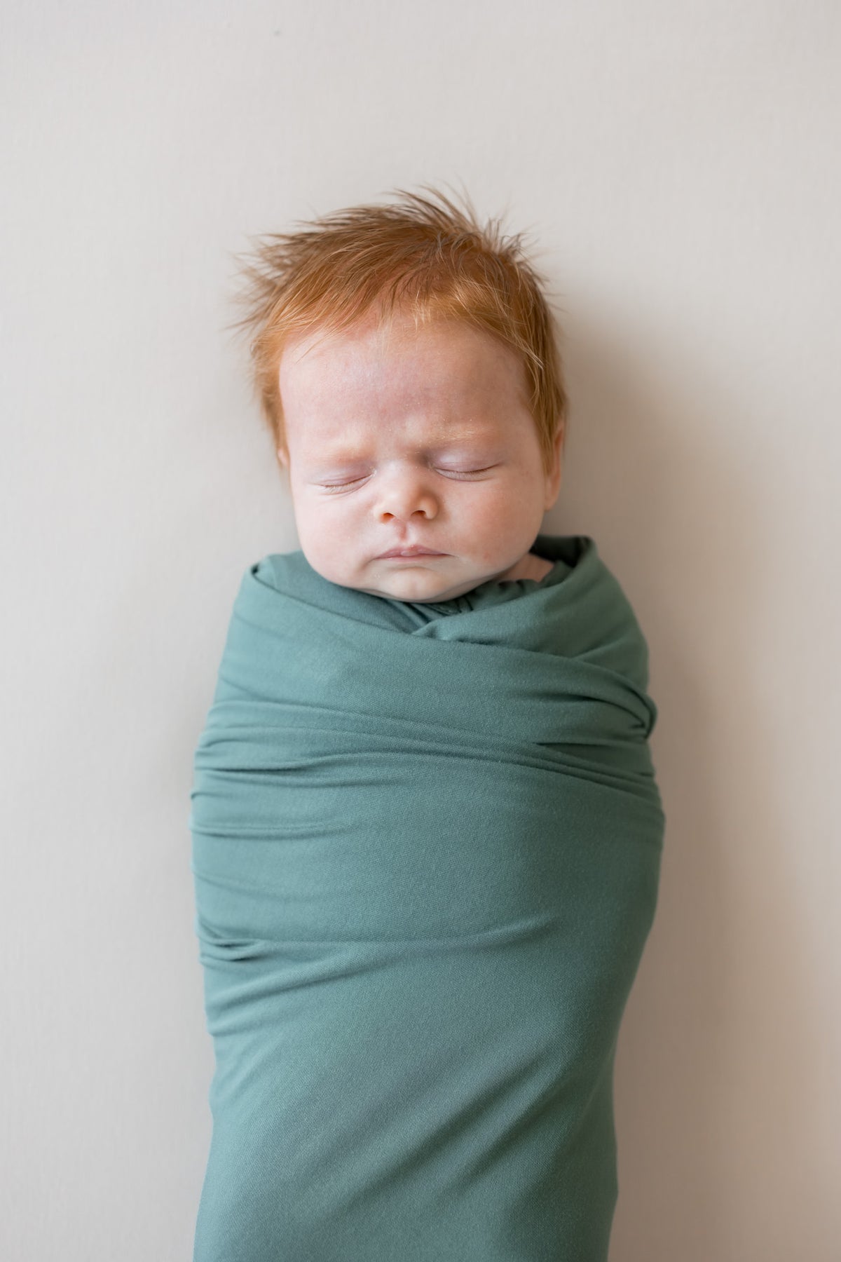 A sleeping newborn baby with red hair, swaddled in a dusty green fabric wrap, standing against a plain light beige wall.
