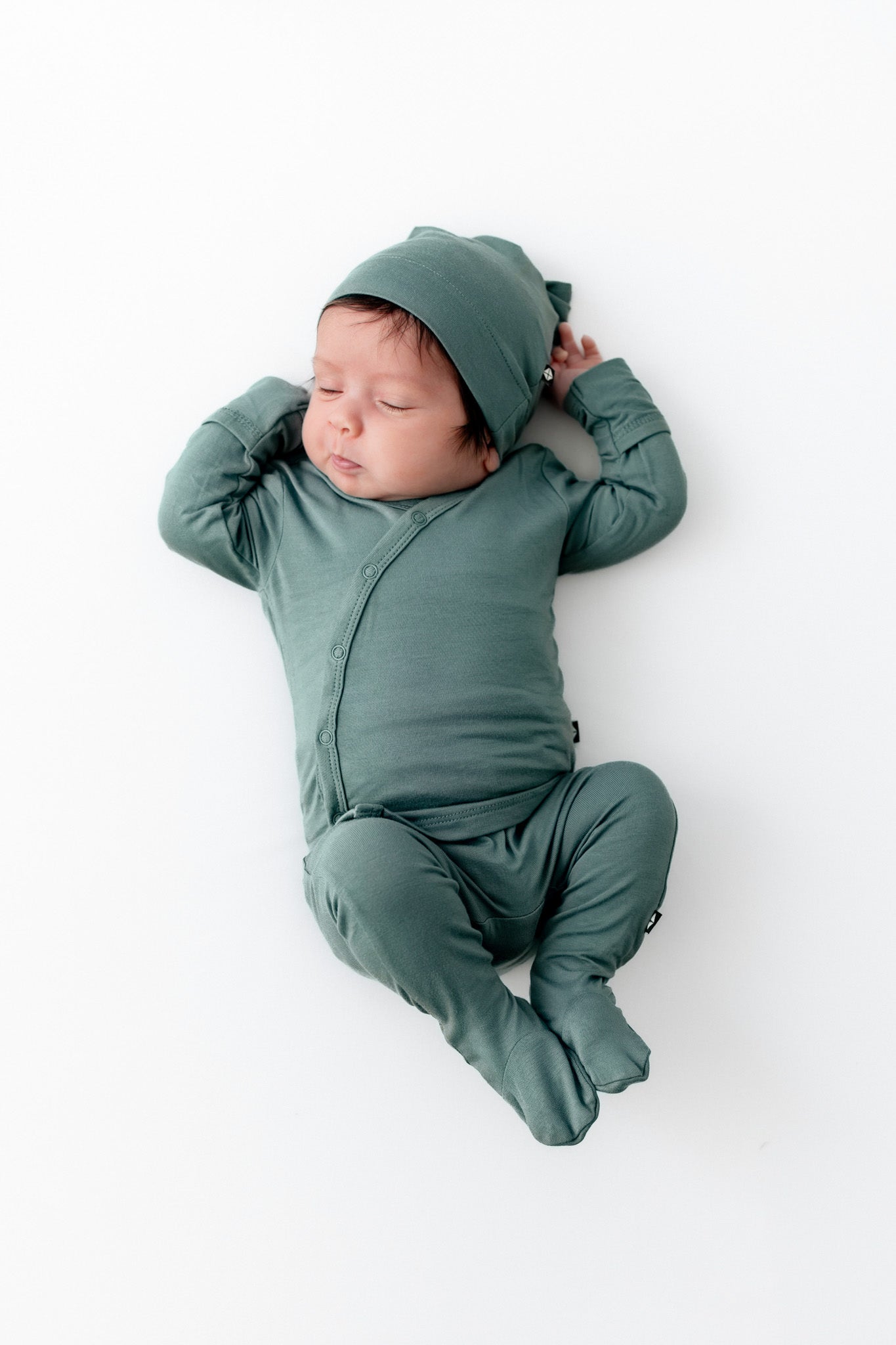 A sleeping newborn baby wearing a matching sage green footed take me home set and cap, lying on their back against a clean white background.