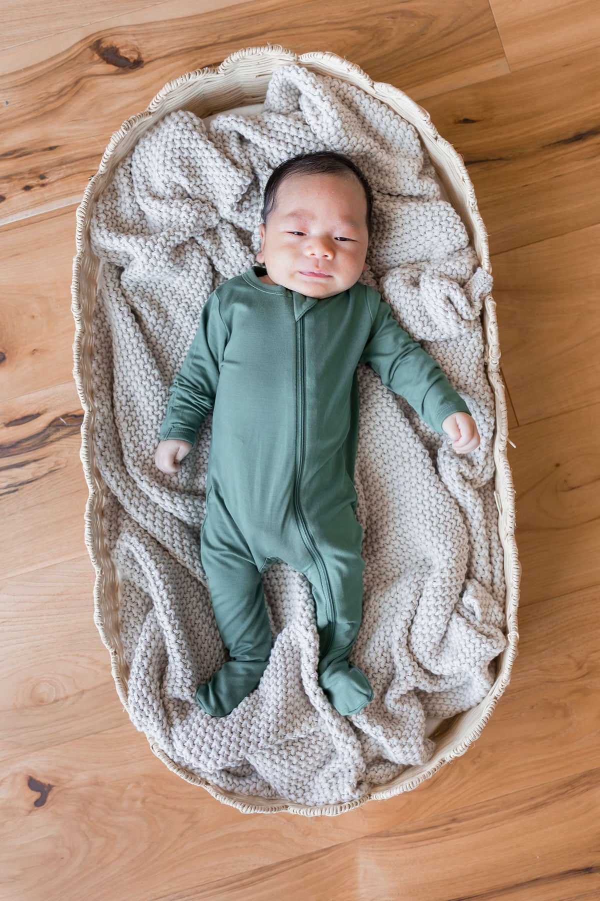 A newborn baby wearing a sage green footed zip-up sleeper, lying on a textured cream knit blanket inside a woven oval bassinet on a wooden floor.