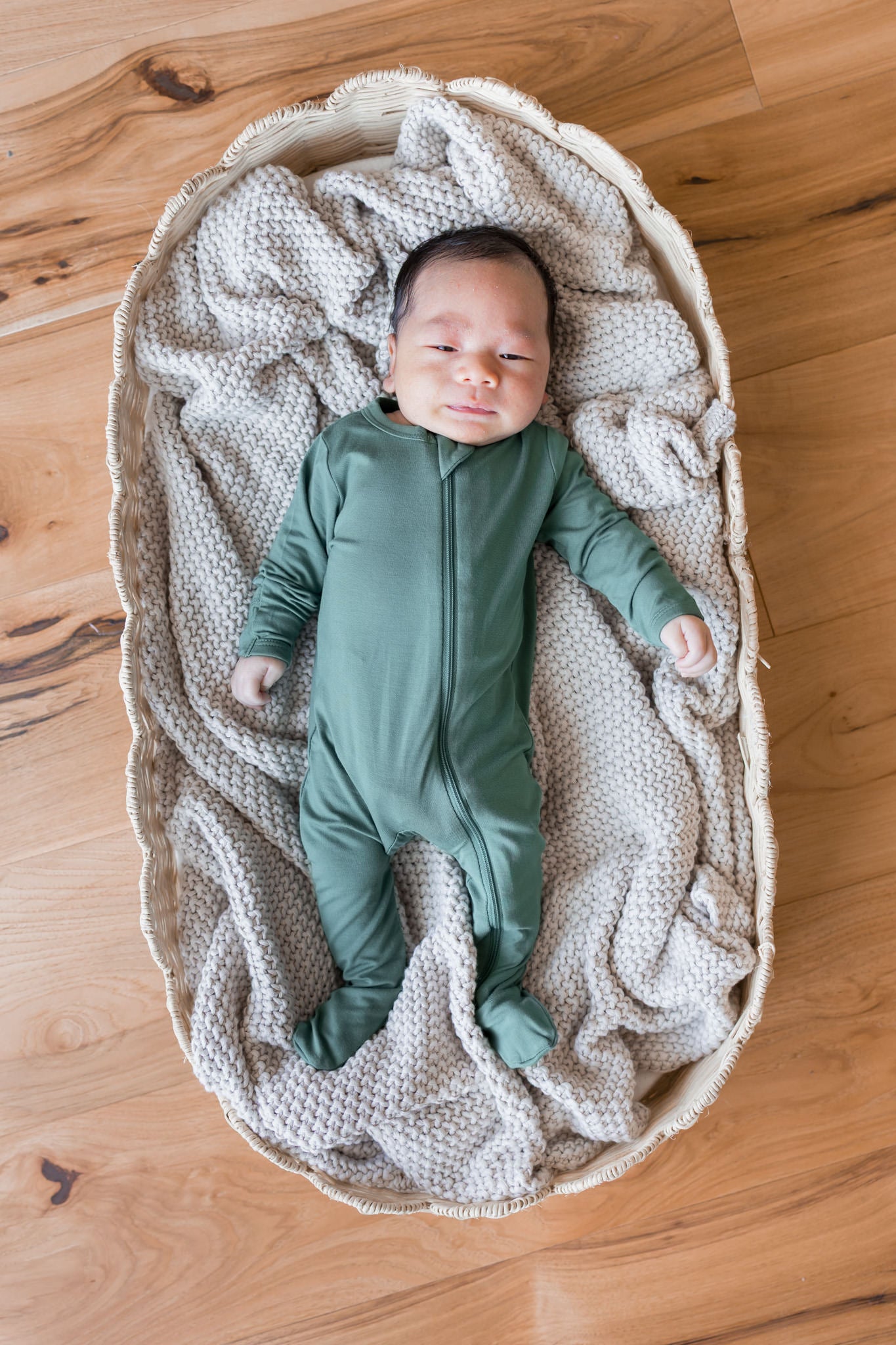 A newborn baby wearing a sage green footed zip-up sleeper, lying on a textured cream knit blanket inside a woven oval bassinet on a wooden floor.