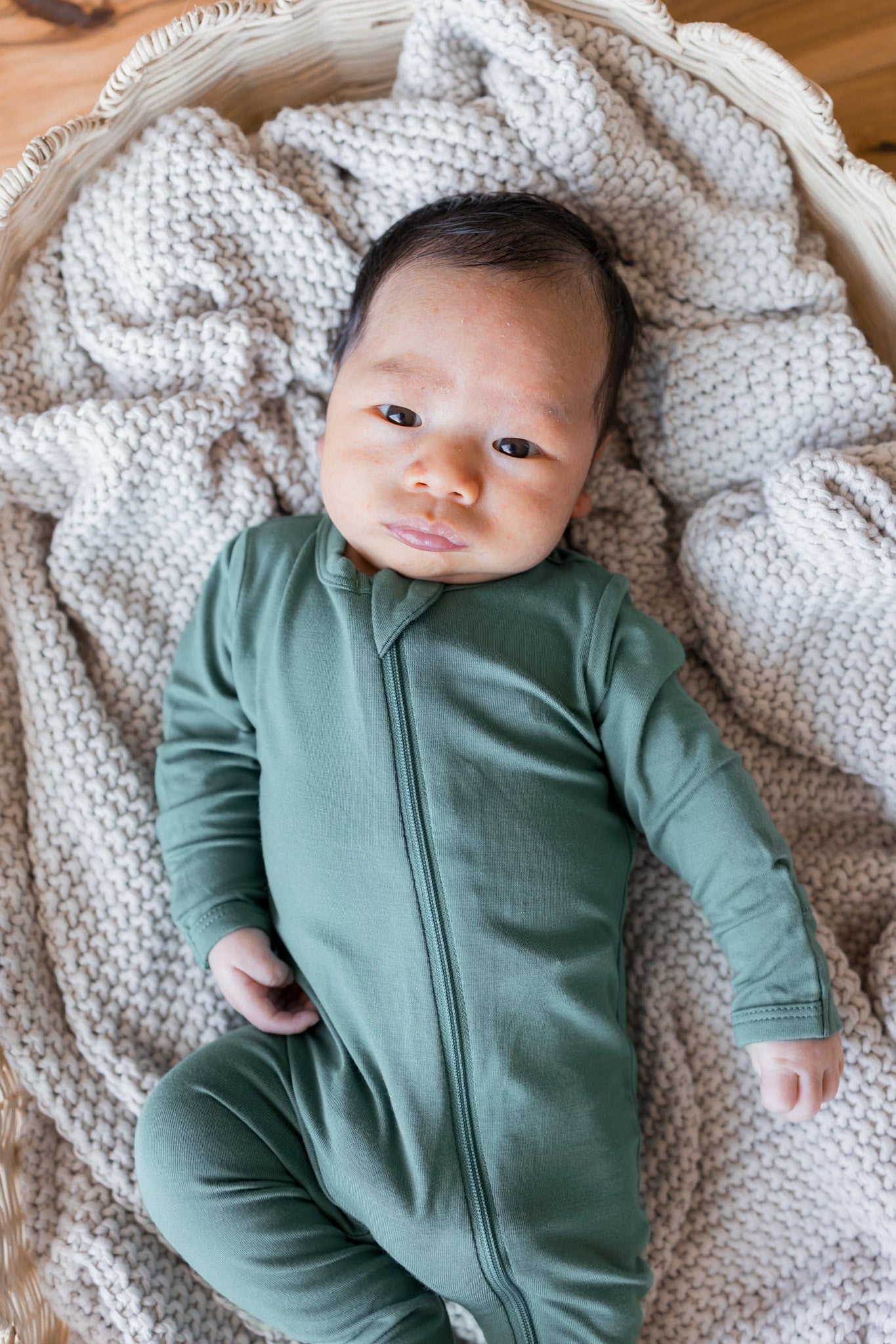 A newborn baby wearing a sage green long-sleeve zip-up onesie, lying on a chunky knit cream blanket inside a woven basket.
