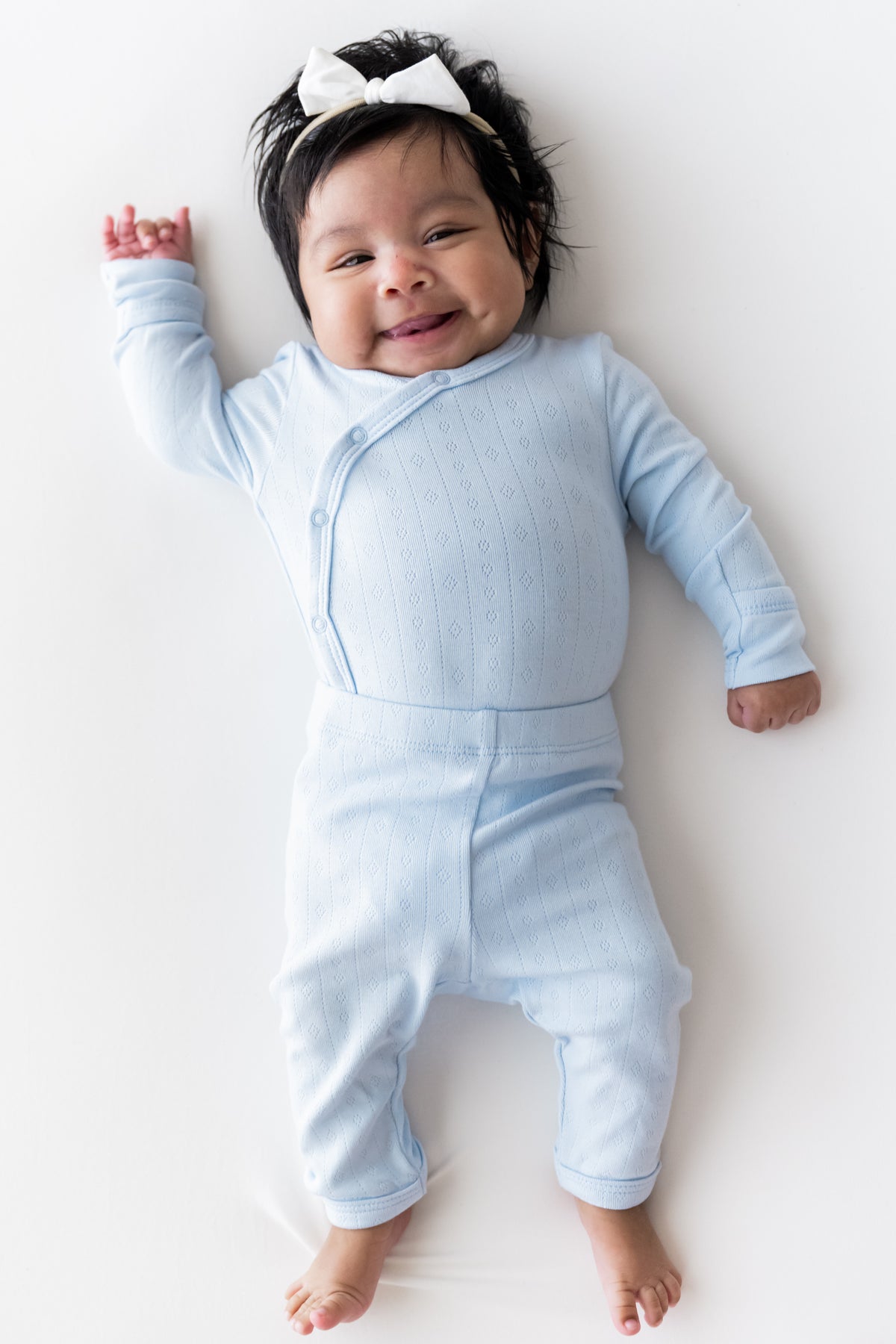 A smiling baby lying on their back wearing a light blue ribbed long-sleeve footless kimono set and a white bow headband, photographed against a clean white background.