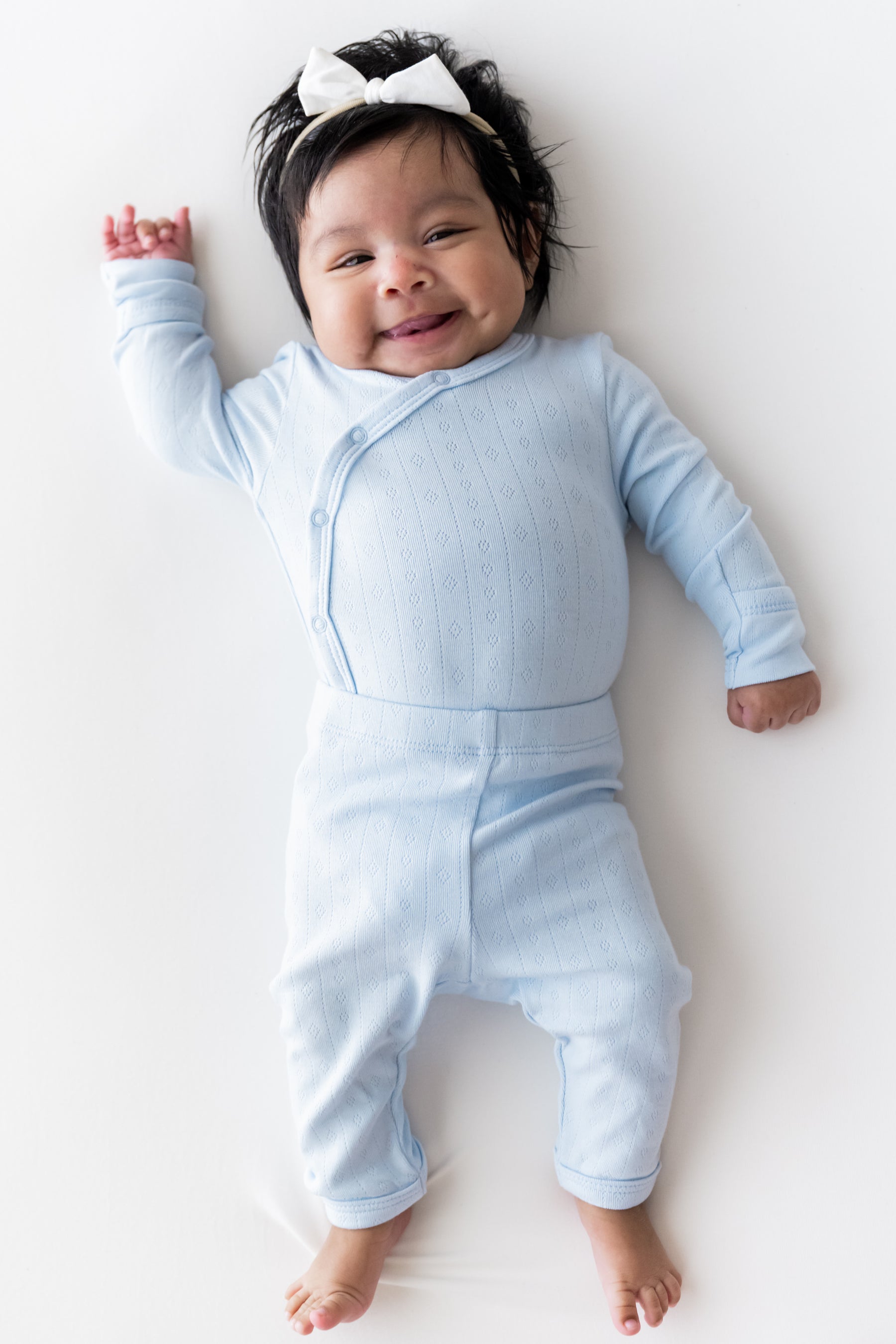 A smiling baby lying on their back wearing a light blue ribbed long-sleeve footless kimono set and a white bow headband, photographed against a clean white background.