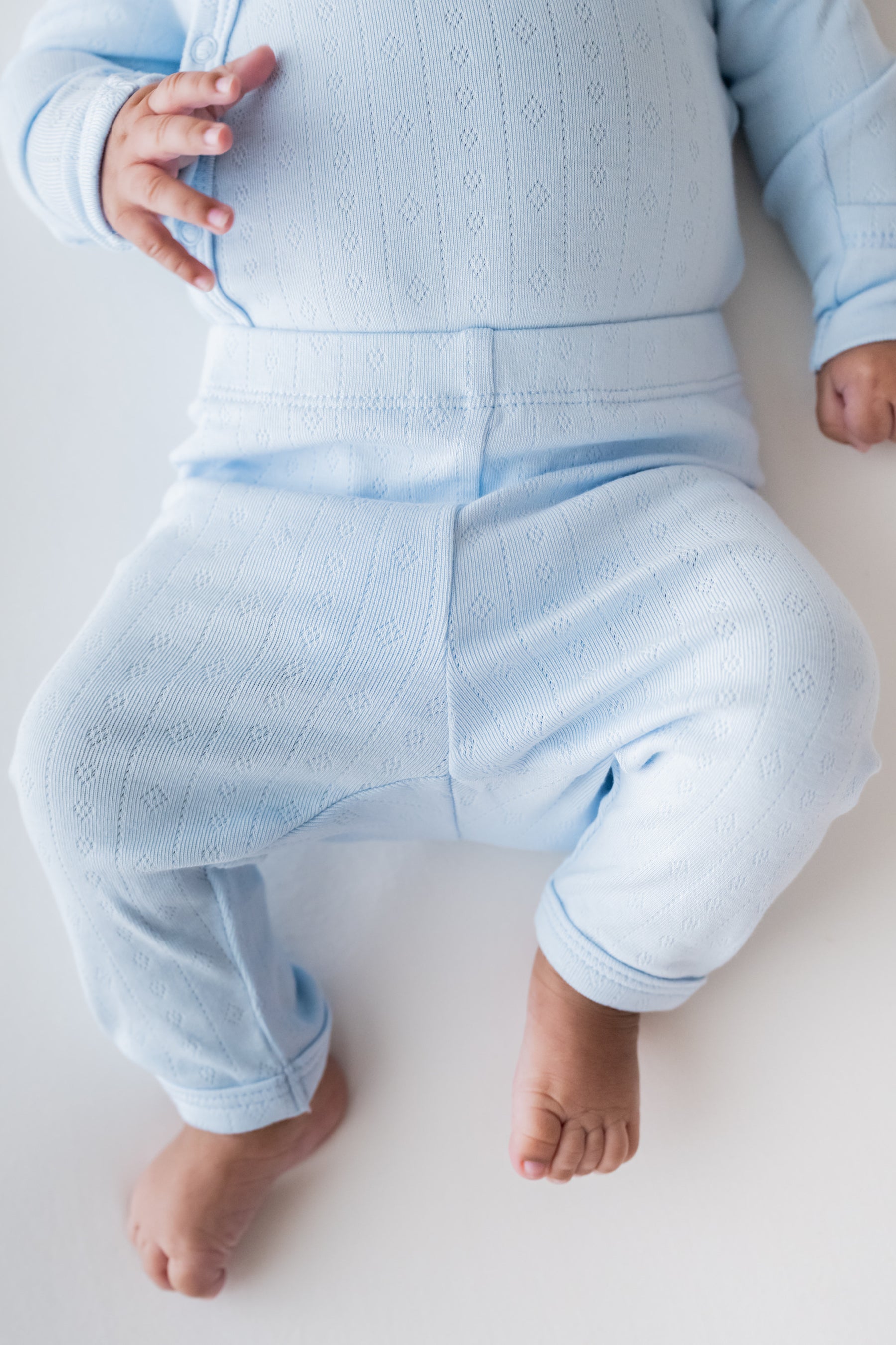 A close-up of a baby wearing a light blue knit two-piece outfit, featuring a long-sleeve top and matching pants, laid out on a white background.