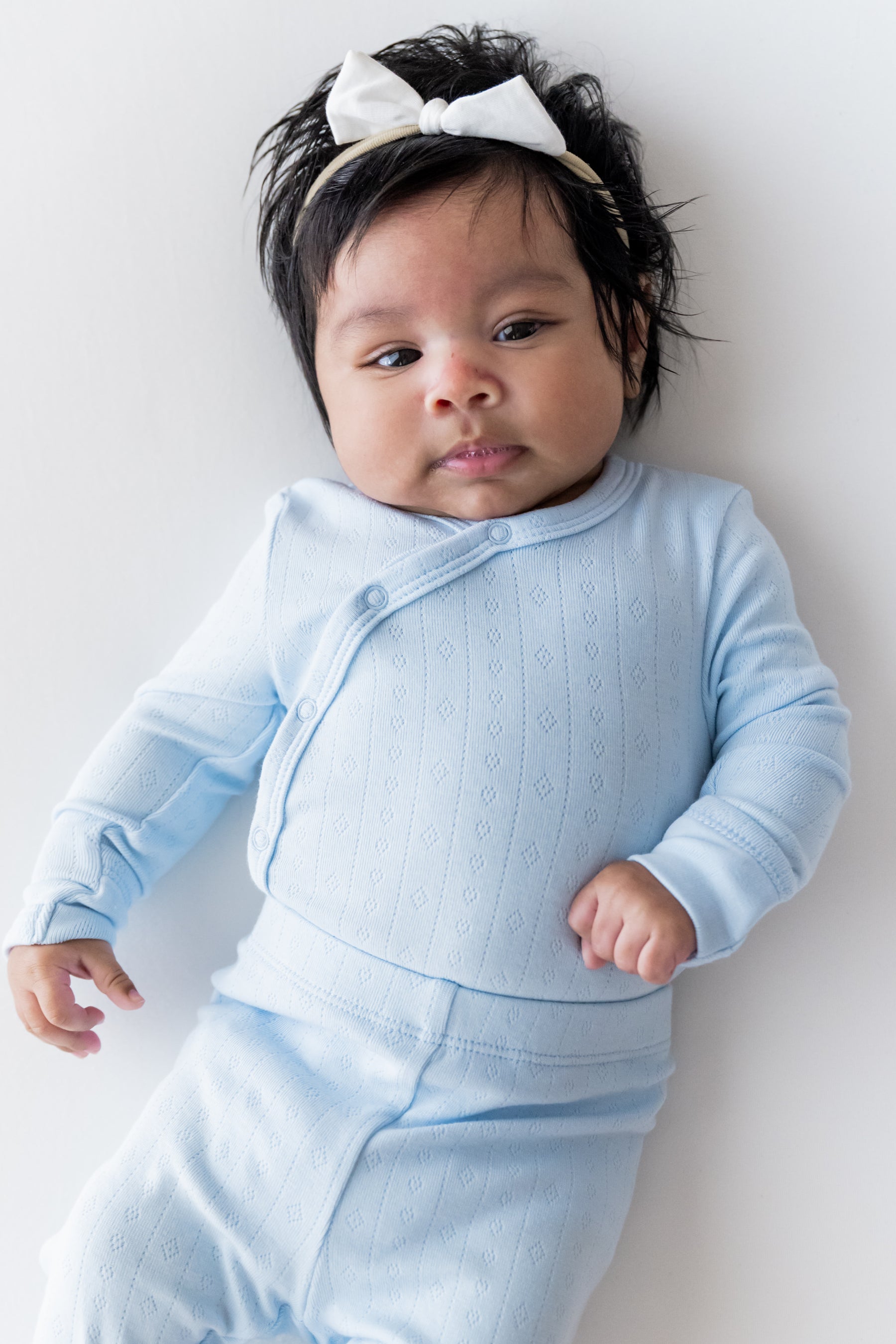 A baby wearing a light blue long-sleeve kimono bodysuit and matching pants, with a white bow headband, lying on a light gray background.