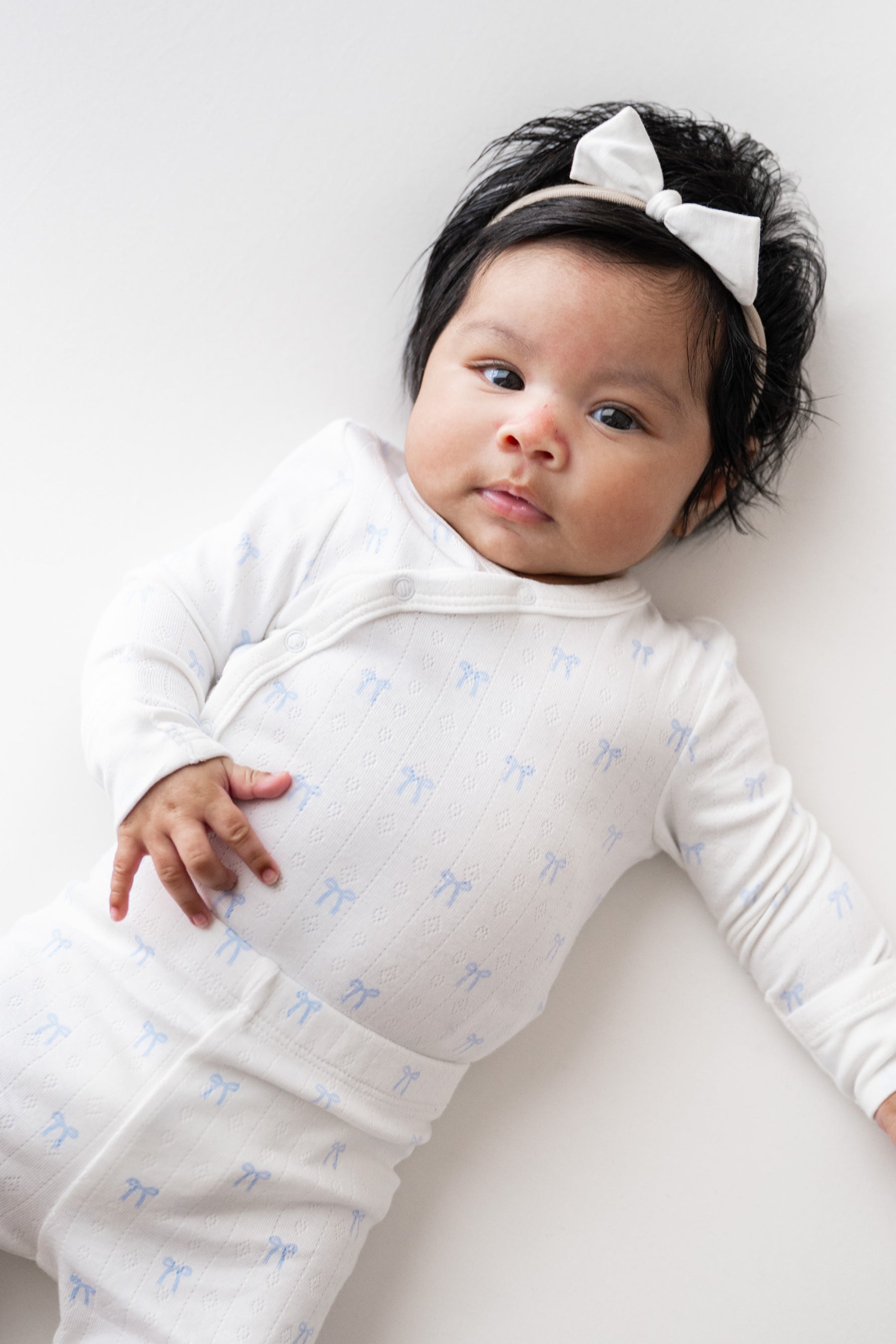 A baby lying on a light background wearing a white long-sleeve bodysuit with a small blue bow pattern and a matching white bow headband.