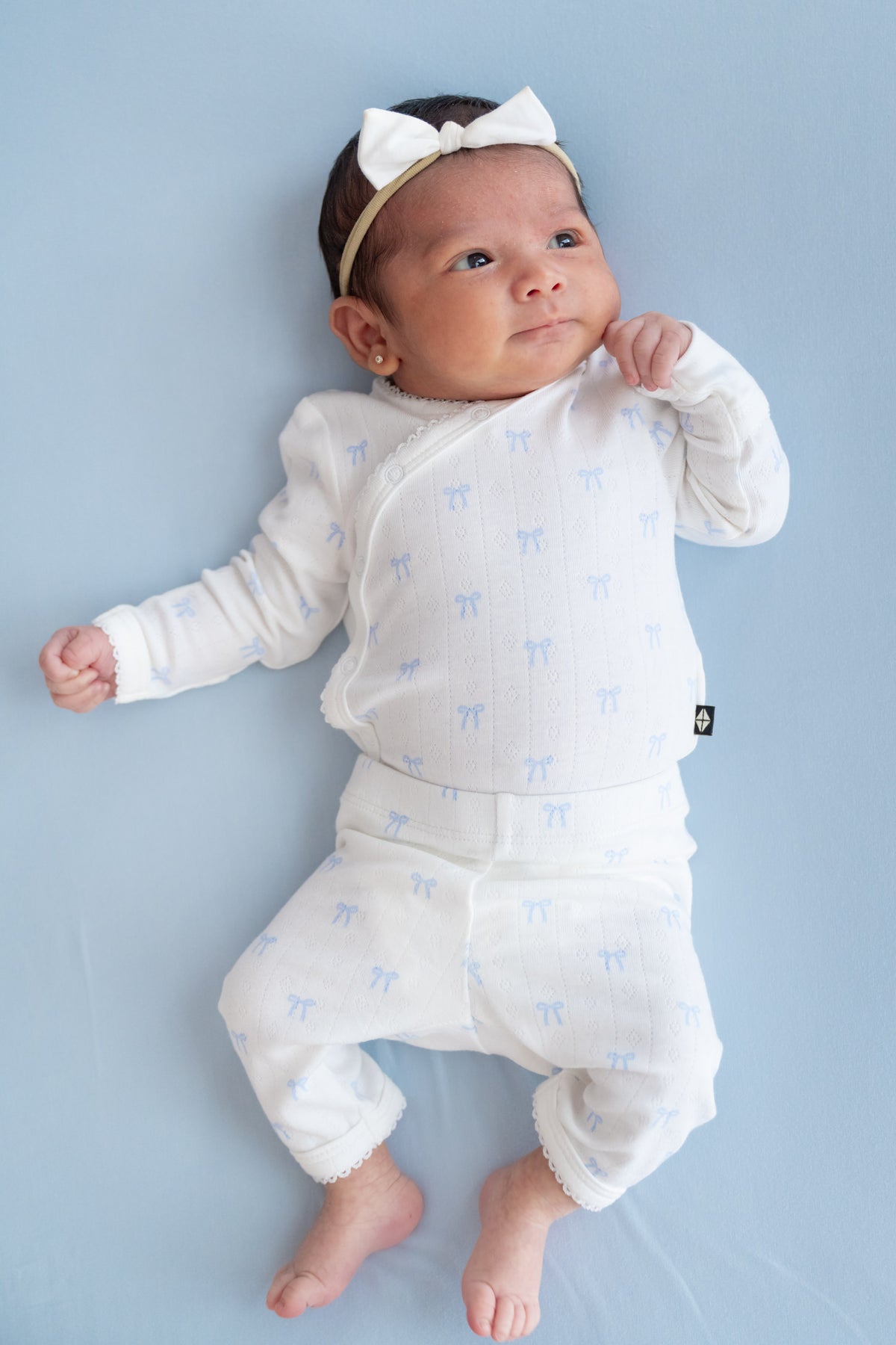 A baby lying on a light blue background, wearing a white long-sleeve kimono and pant with a light blue bow pattern, a matching white headband with a bow, and small gold hoop earrings.