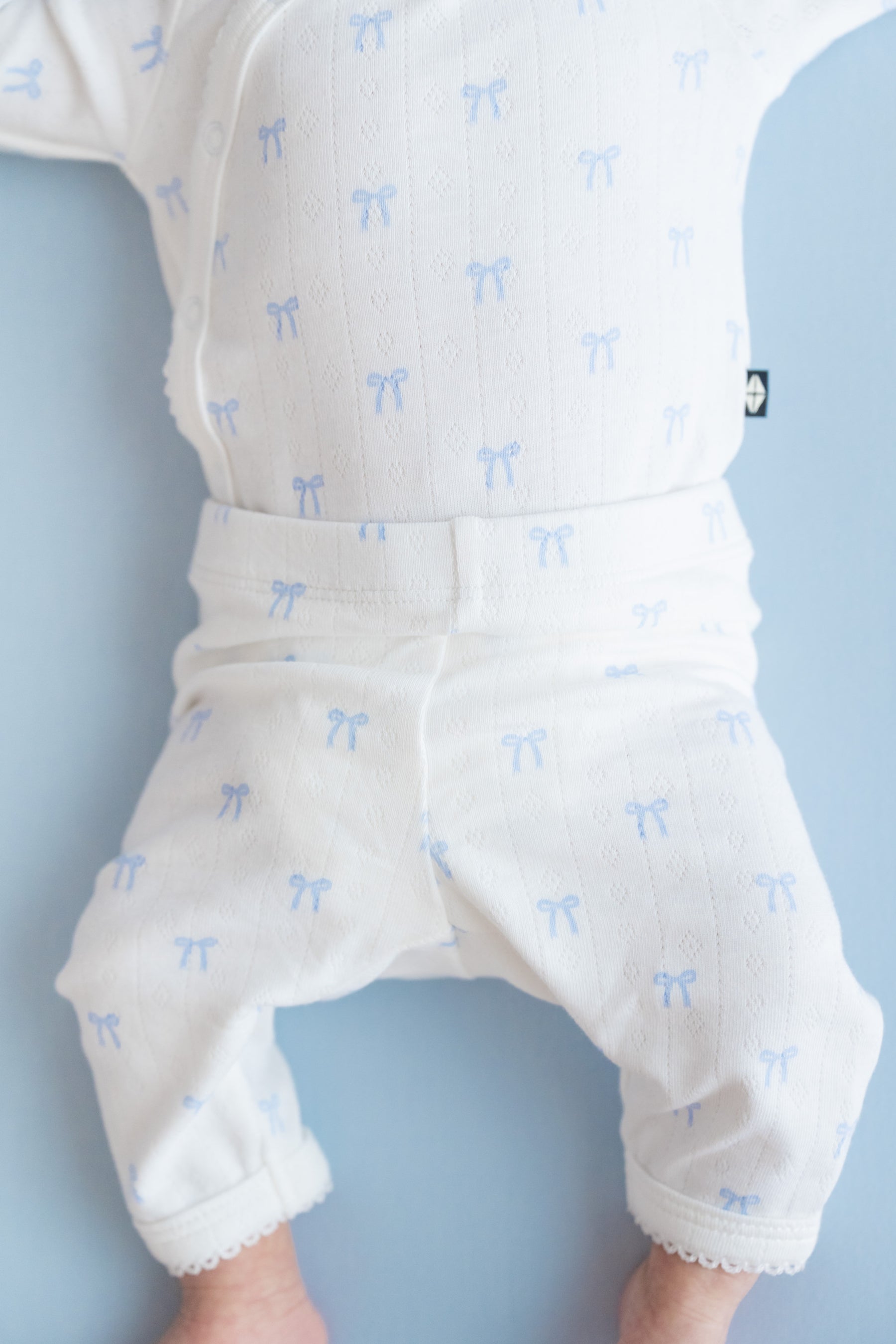 A close-up of a baby wearing a white pointelle kimono and matching pants with a light blue bow pattern, lying on a light blue background.