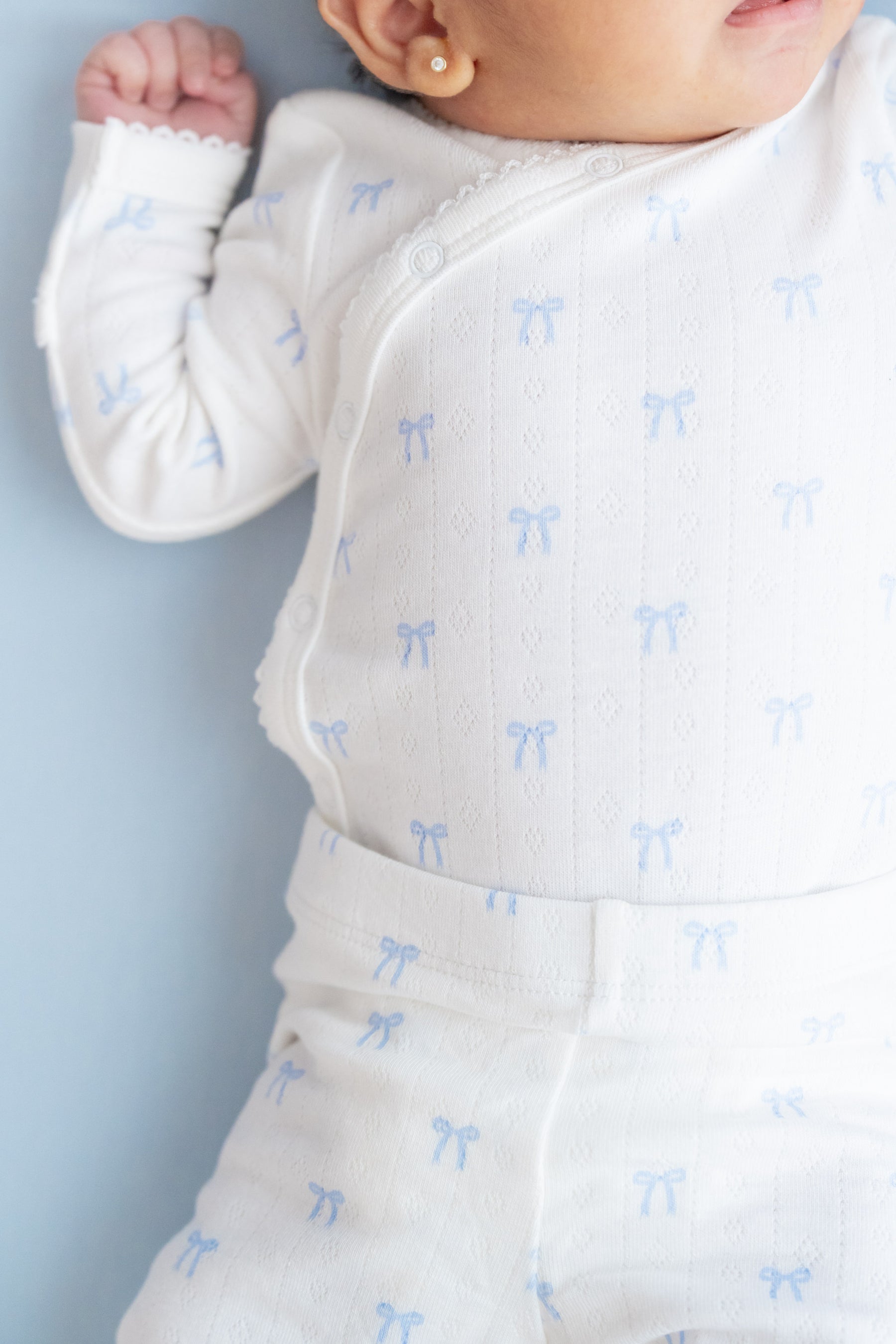 A close-up of a baby wearing a white long-sleeve bodysuit with a repeating light blue bow pattern, lying on a light blue background.
