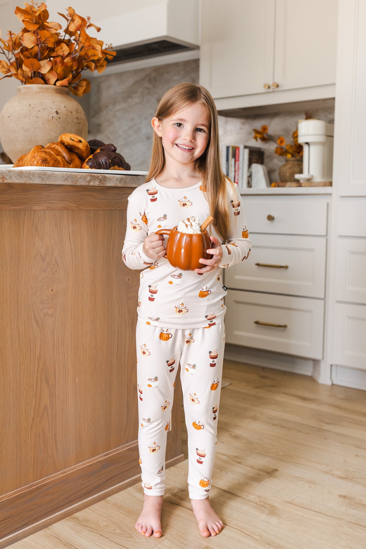 Girl standing in kitchen wearing Long Sleeve Pajamas with Pumpkin Spice Latte print holding a spice latte cup.
